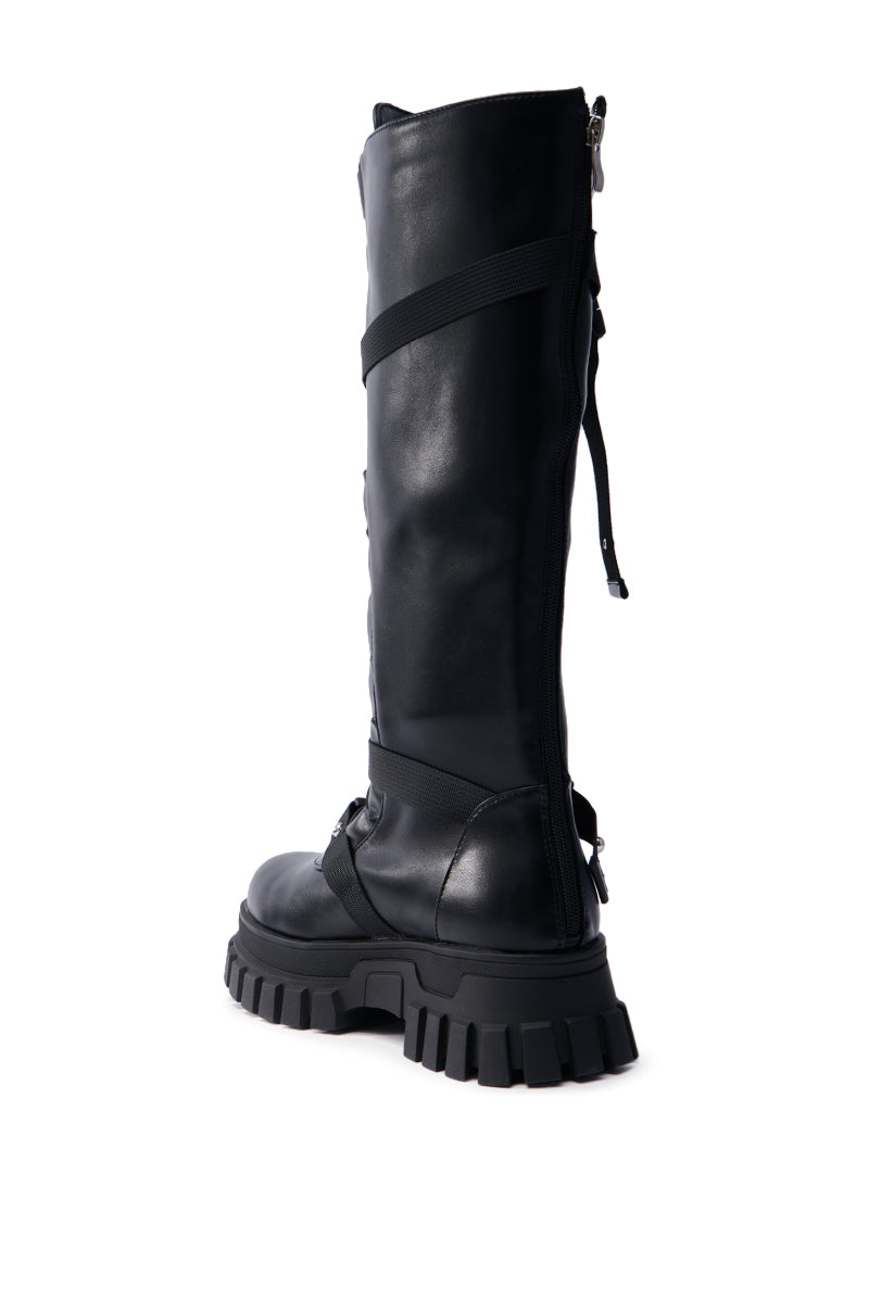 The AZALEA WANG MYRON BLACK BOOT is a black knee-high faux leather combat boot featuring rugged chunky soles, a side zipper, and strap details around the calf and heel for a sleek, edgy punk look. Angled to highlight its back and side profile.