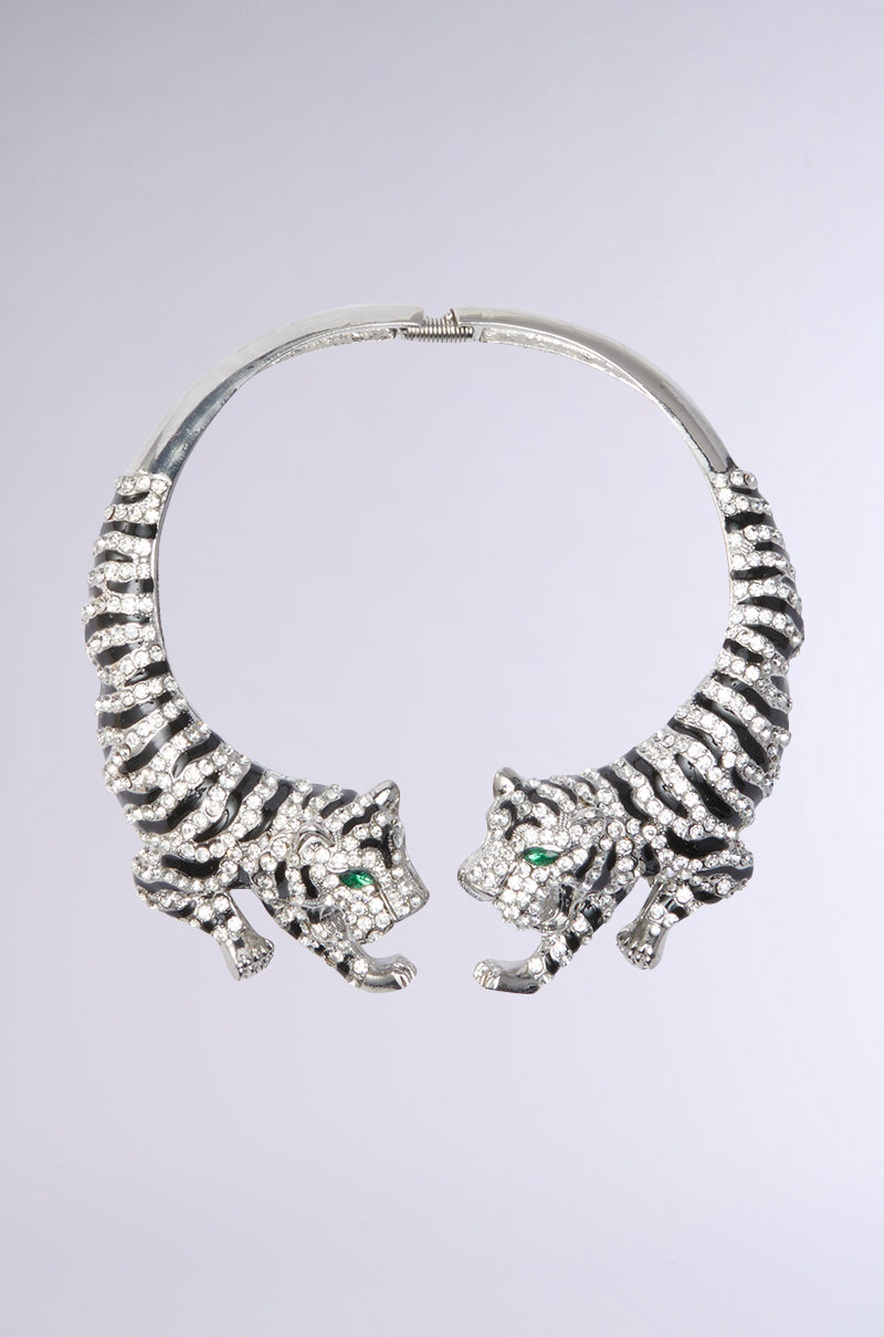 The ON THE PROWL CHOKER by AKIRA Label features two silver tigers with green eyes and black stripes facing each other. Adorned with small gemstones, this elegant piece contrasts vividly against its shimmering backdrop, embodying the essence of a Rhinestone Tiger. 1