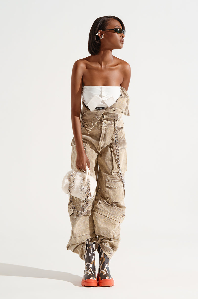 A person wearing the NO ONE LIKE ME CARGO DENIM JUMPSUIT, which features a strapless white top and tan wide-leg silhouette cargo pants with chains, holds a fluffy white bag while posing confidently. They accessorize with sunglasses and multicolored boots featuring a camouflage pattern against a plain white background.