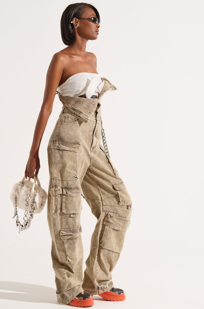 A fashionable individual poses against a white background, wearing the NO ONE LIKE ME CARGO DENIM JUMPSUIT, featuring an oversized stone-washed design with a strapless top. They hold a furry handbag and wear dark sunglasses, with orange-soled black shoes adding a bold touch to the outfit.