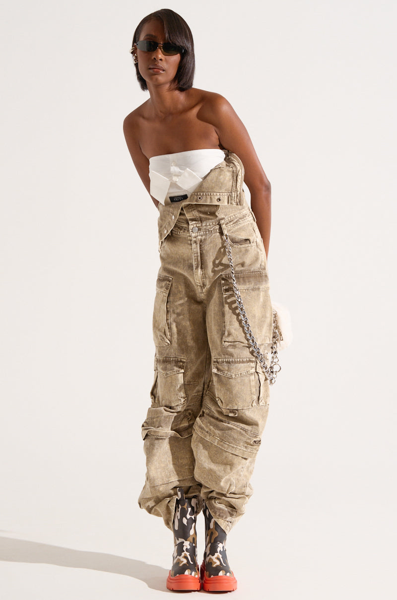 A person wearing sunglasses poses in a fashion-forward outfit, featuring a white strapless top, the NO ONE LIKE ME CARGO DENIM JUMPSUIT with multiple pockets and a hanging chain, and black and white patterned boots with orange soles. The wide leg silhouette adds an extra touch of modern chic to the look. The background is plain white.