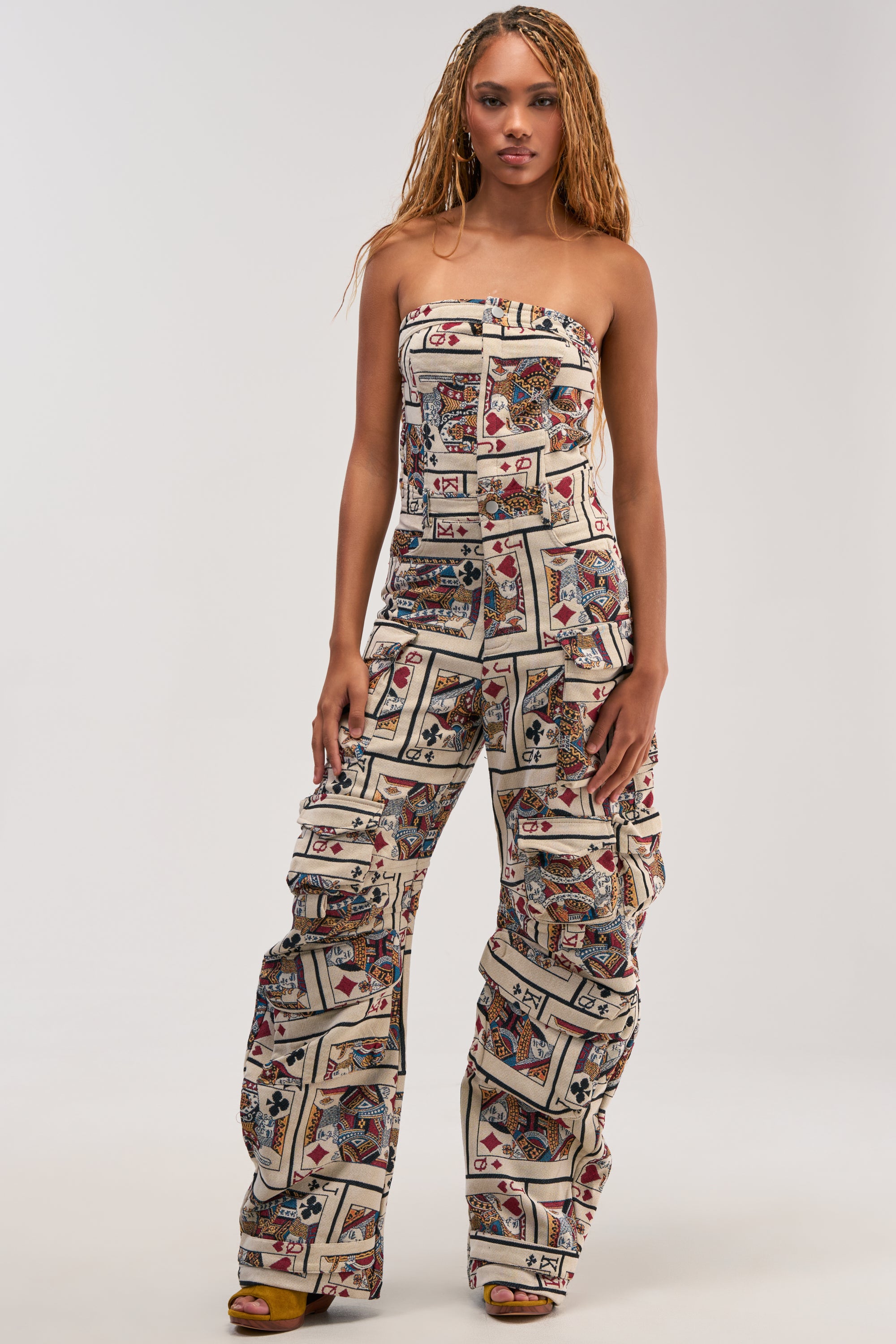 A woman models the NO ONE LIKE ME Card Print Cargo Jumpsuit, featuring a colorful patchwork design and wide-leg pants, standing against a plain white background.