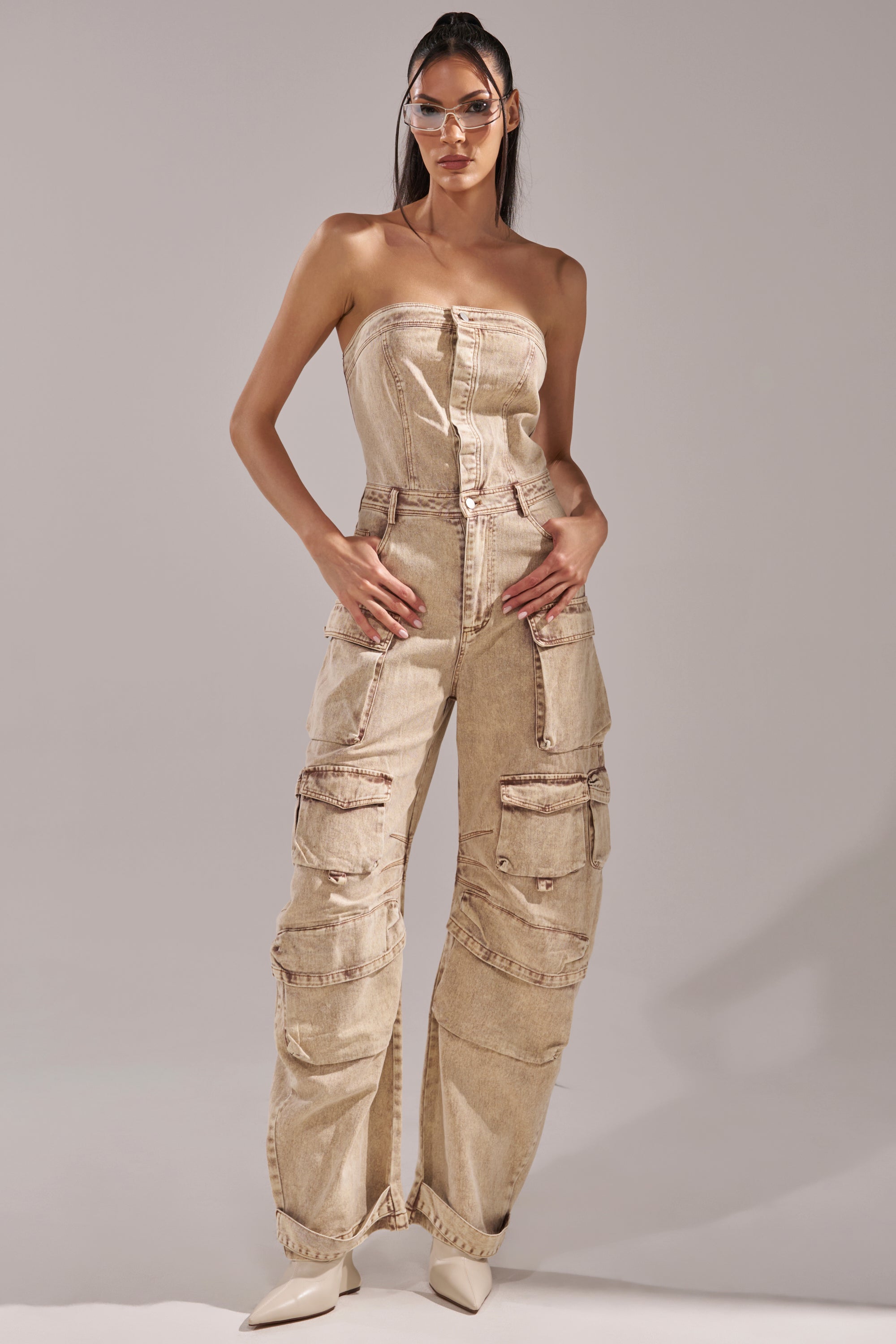 A woman in clear glasses and the JESSICA DENIM CARGO JUMPSUIT with multiple cargo pockets poses confidently in white pointed-toe heels against a plain background. 0