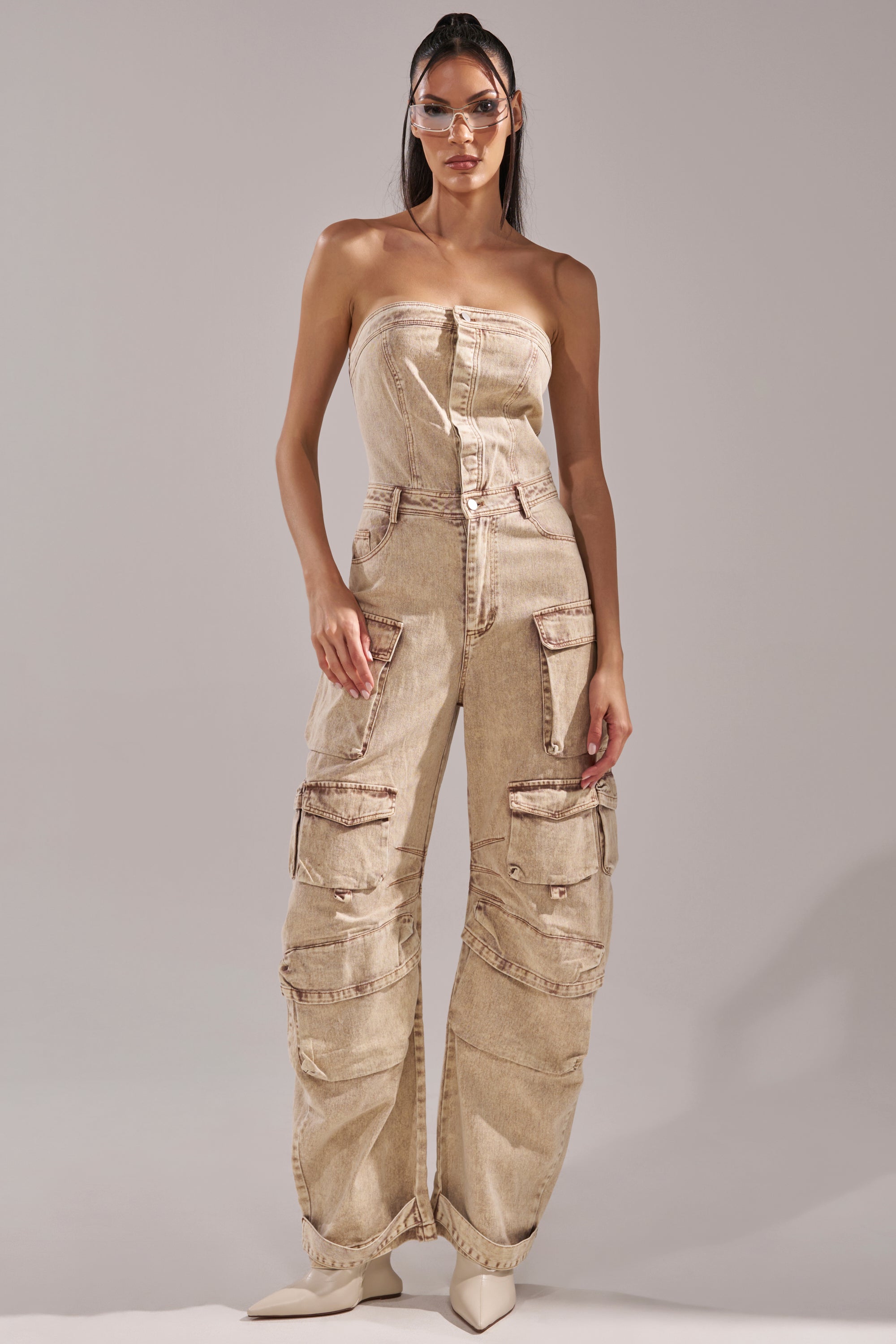 A woman wears the JESSICA DENIM CARGO JUMPSUIT, a strapless denim piece with multiple pockets and seams, styled with pointed-toe boots and clear visor glasses against a plain neutral background. 1