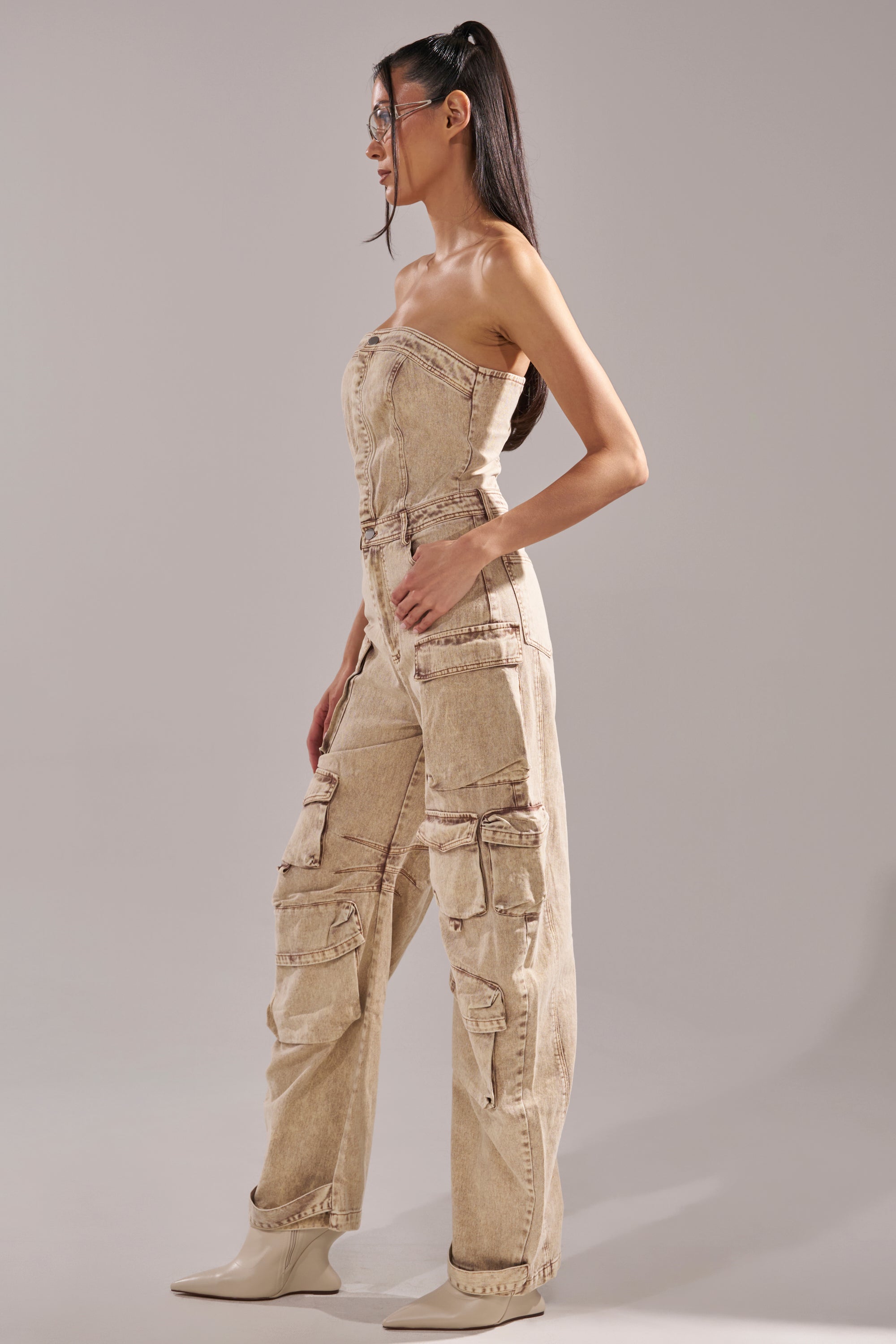 A woman in profile wears the JESSICA DENIM CARGO JUMPSUIT, featuring a strapless design with multiple pockets and seam details. She has long dark hair in a high ponytail, beige pointed boots, clear glasses, and stands against a plain gray background. 3