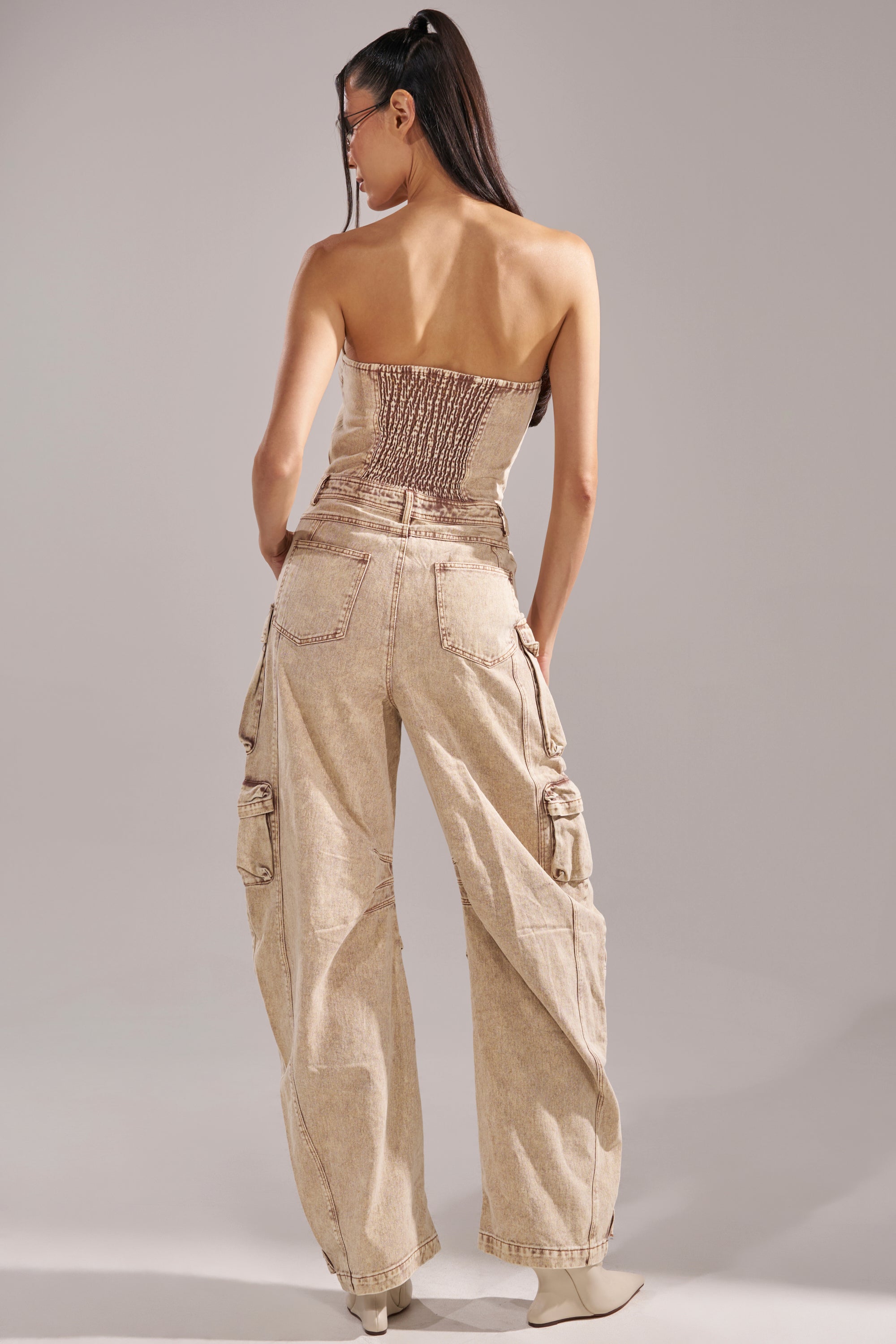 A woman faces away in the JESSICA DENIM CARGO JUMPSUIT, featuring a strapless smocked back and loose cargo pockets. She pairs it with light shoes, has straight long hair, and wears sunglasses. 4