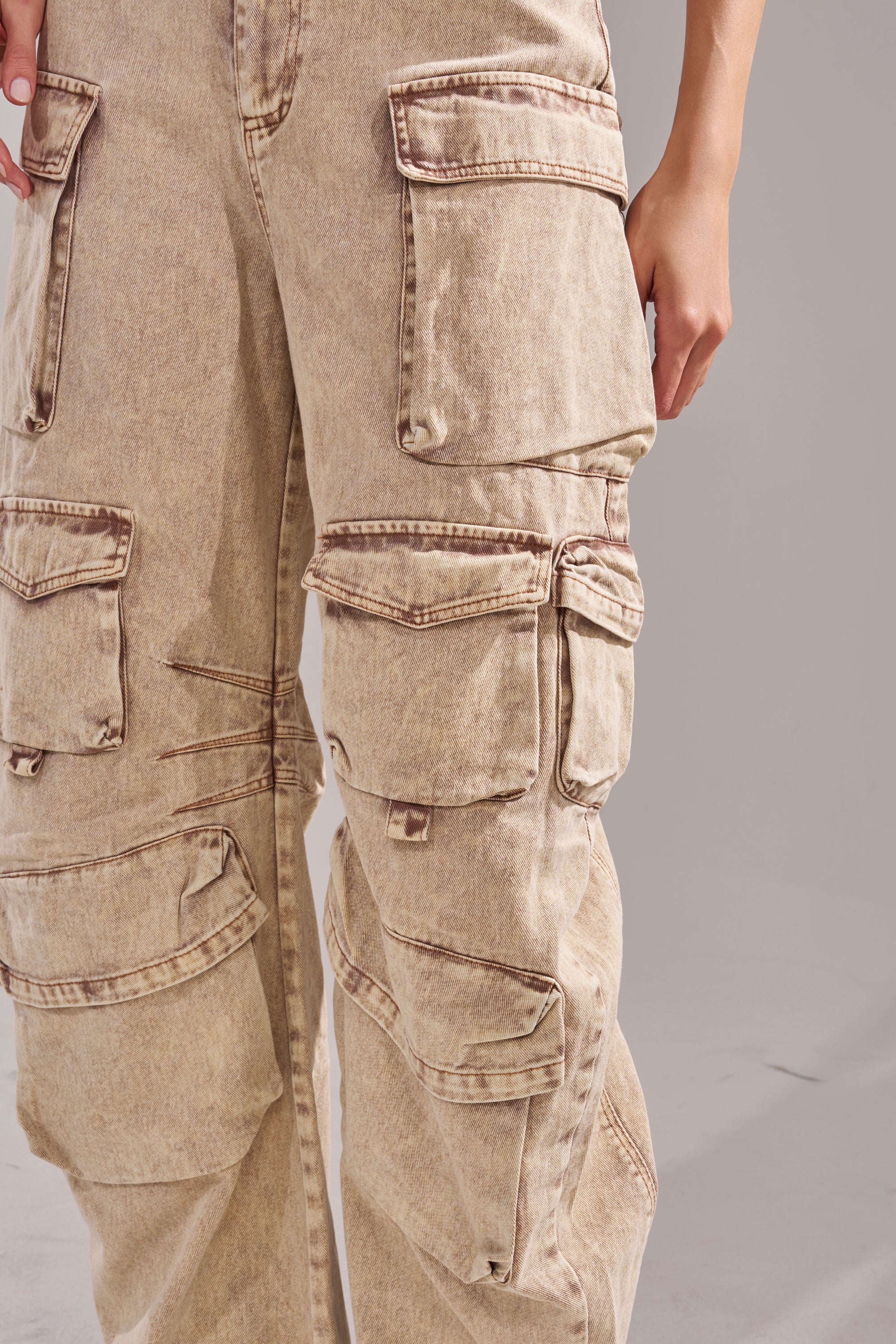 A person in the JESSICA DENIM CARGO JUMPSUIT, featuring multiple large pockets and an AKIRA Label-inspired design, stands against a plain light background, with only the lower torso and one hand visible. 5