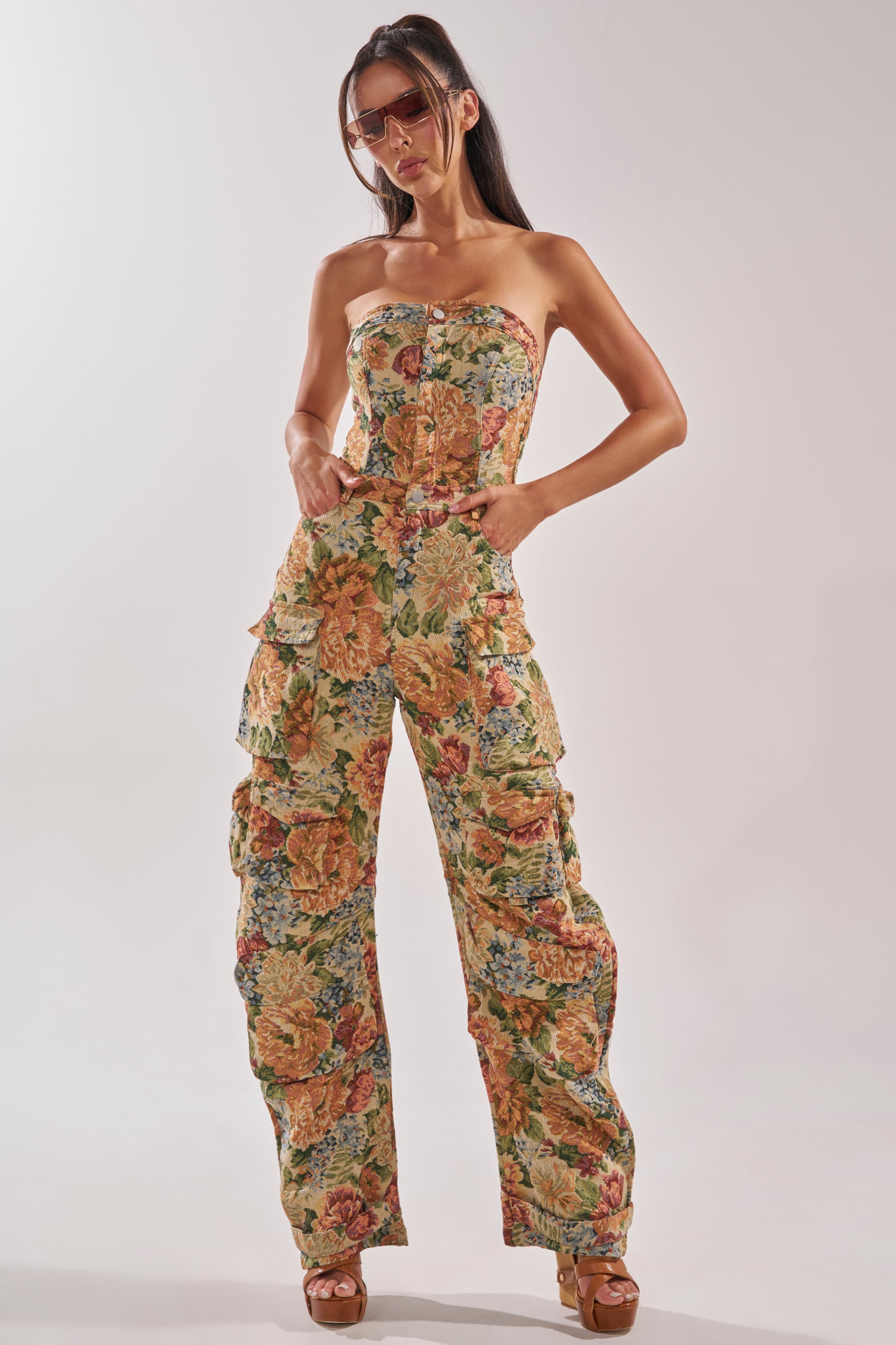 A woman wears the IN HER ELEMENT FLORAL WOVEN JUMPSUIT IN BROWN MULTI—strapless with a colorful floral print and ruffle leg details—paired with brown platform heels and large sunglasses, standing hands in pockets against a plain backdrop. 0