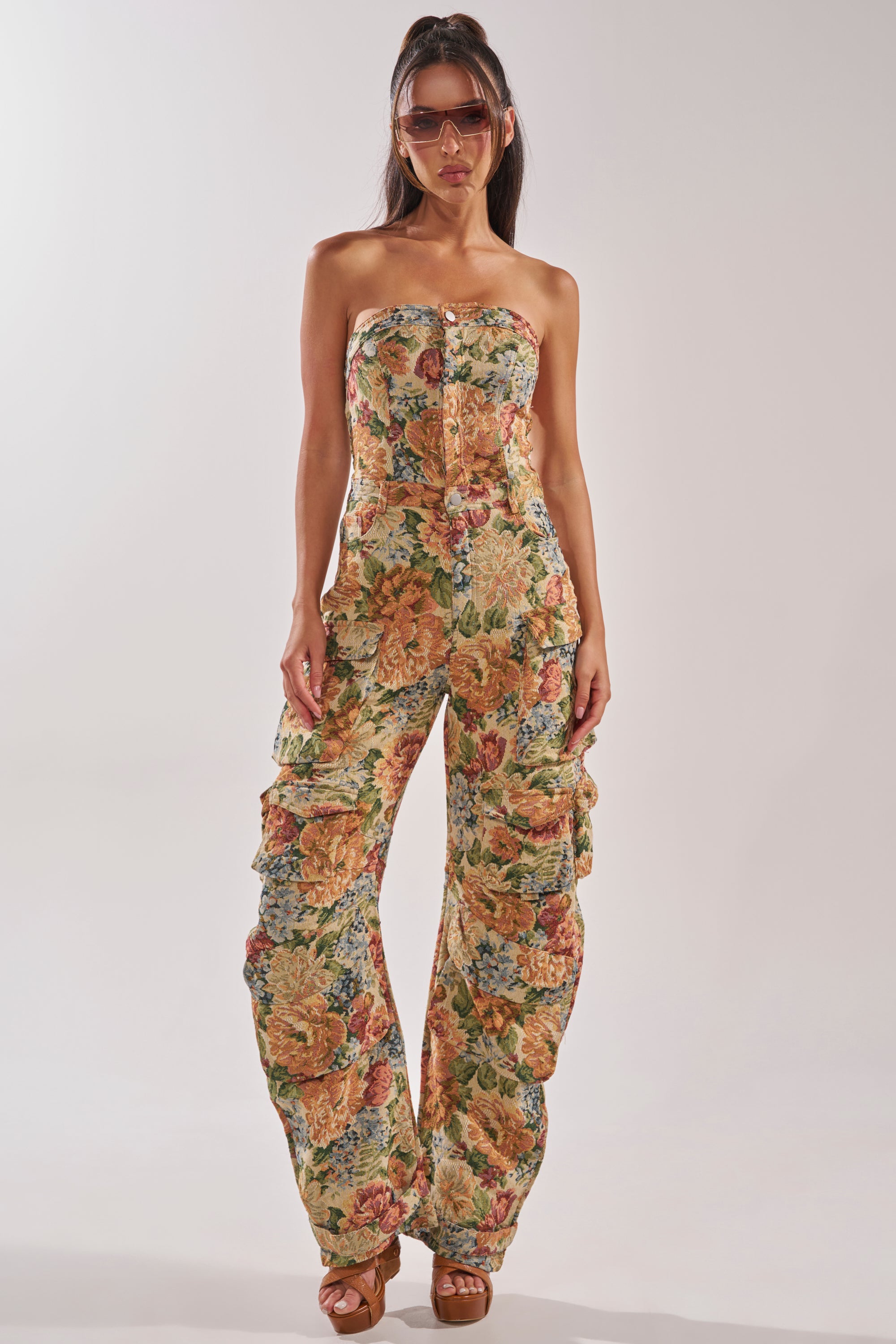 A woman stands against a light background wearing the IN HER ELEMENT FLORAL WOVEN JUMPSUIT IN BROWN MULTI, featuring a strapless floral print and ruched barrel leg, paired with brown platform sandals and oversized sunglasses. 1