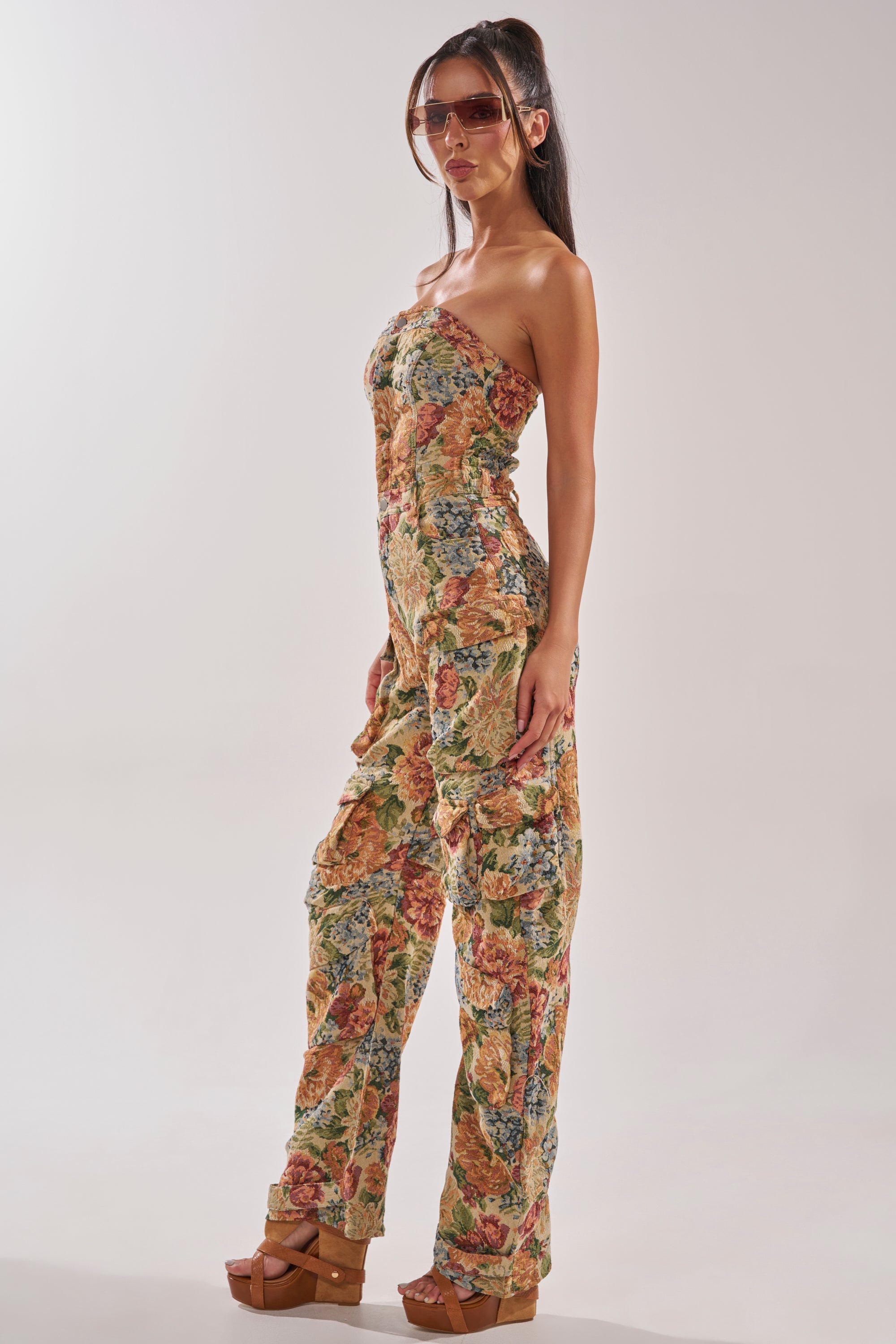 A woman wearing the IN HER ELEMENT FLORAL WOVEN JUMPSUIT IN BROWN MULTI, brown platform sandals, and large sunglasses poses sideways against a plain light background. 2