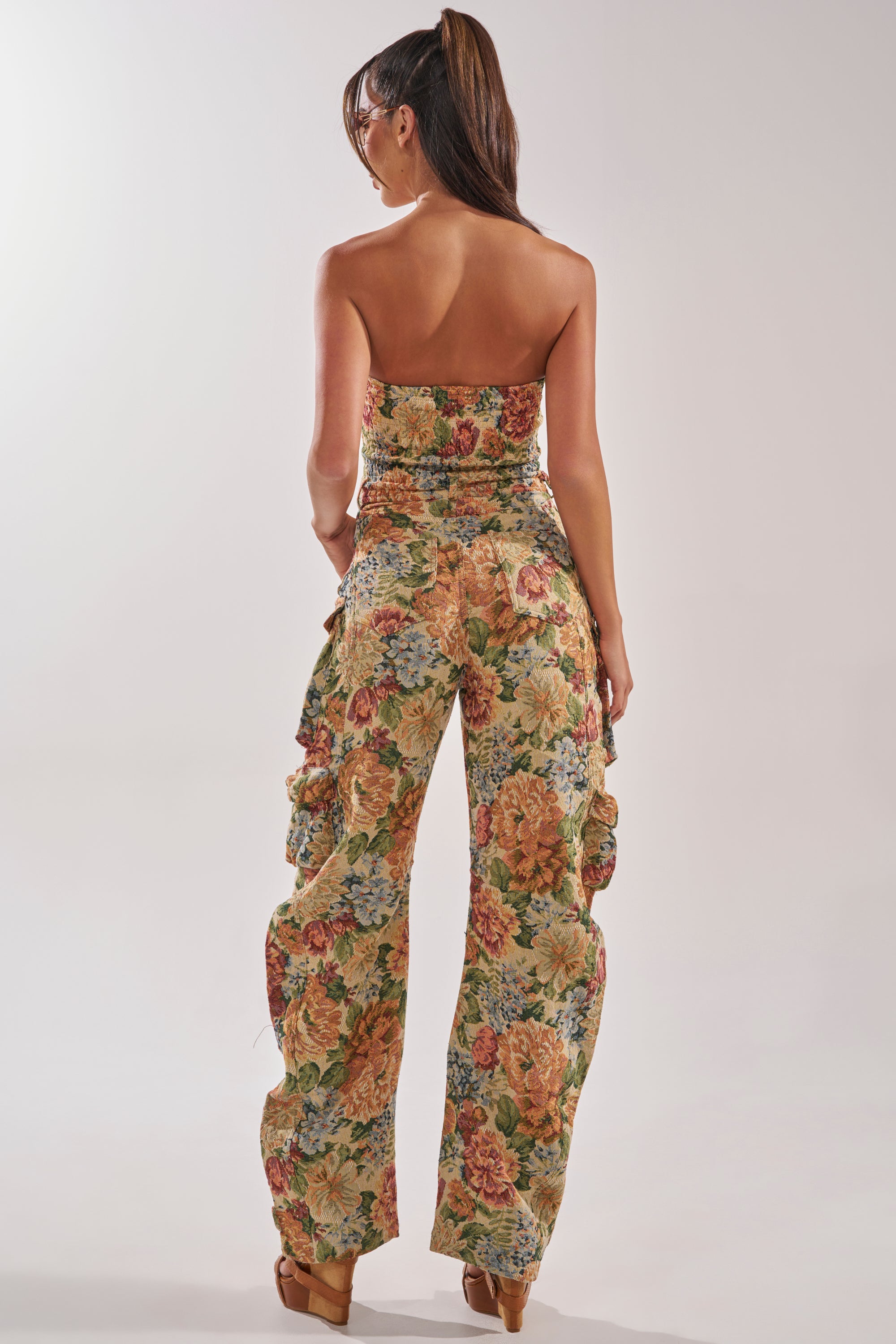 A woman with a high ponytail stands with her back to the camera, wearing the IN HER ELEMENT FLORAL WOVEN JUMPSUIT IN BROWN MULTI, featuring a strapless floral design, wide-leg pants with ruffles, and tan platform shoes. 3