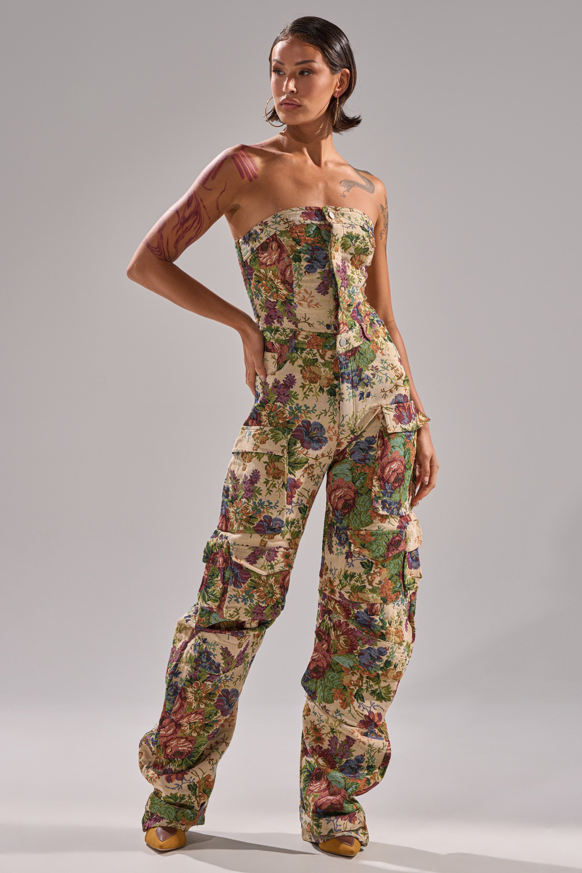 A woman exudes confidence in the IN HER ELEMENT FLORAL WOVEN JUMPSUIT, featuring a strapless design, wide-leg pants, and multiple pockets. With short hair, bold makeup, and statement earrings, she poses against a plain backdrop.