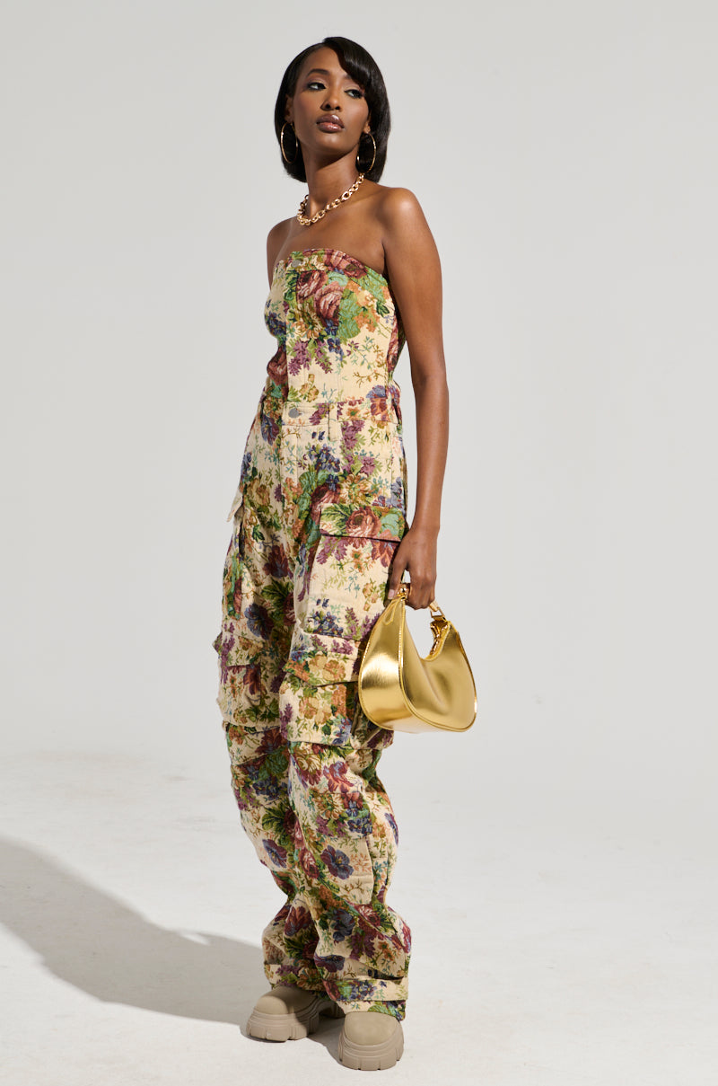 A woman stands confidently in the IN HER ELEMENT FLORAL WOVEN JUMPSUIT, showcasing its vintage floral print and strapless design with a wide-leg silhouette. She accessorizes with a gold purse, hoop earrings, and a necklace while her hair is styled straight. The background is plain and well-lit.