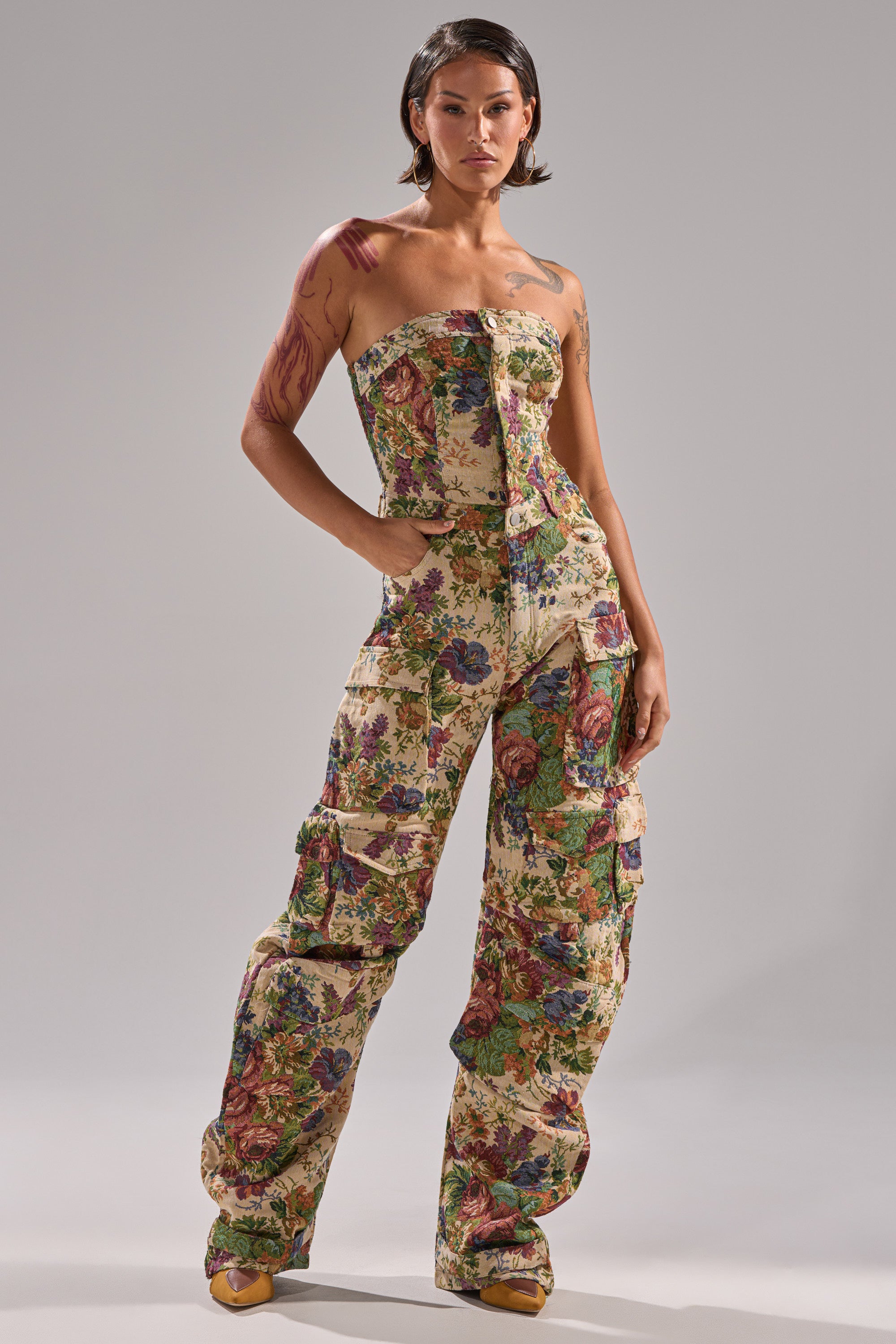 A person stands confidently in the IN HER ELEMENT FLORAL WOVEN JUMPSUIT, featuring a strapless design, cargo pockets, and wide legs. With short hair and visible tattoos on one arm, they pose with hands in pockets against a light gray background.