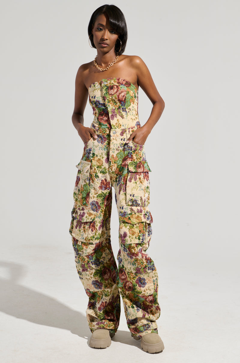 A person poses in the IN HER ELEMENT FLORAL WOVEN JUMPSUIT, showcasing its vintage floral print with a wide-leg silhouette and multiple large pockets. They have their hands in the pockets and are accessorized with a necklace. The jumpsuit features colorful flowers against a plain gray background.
