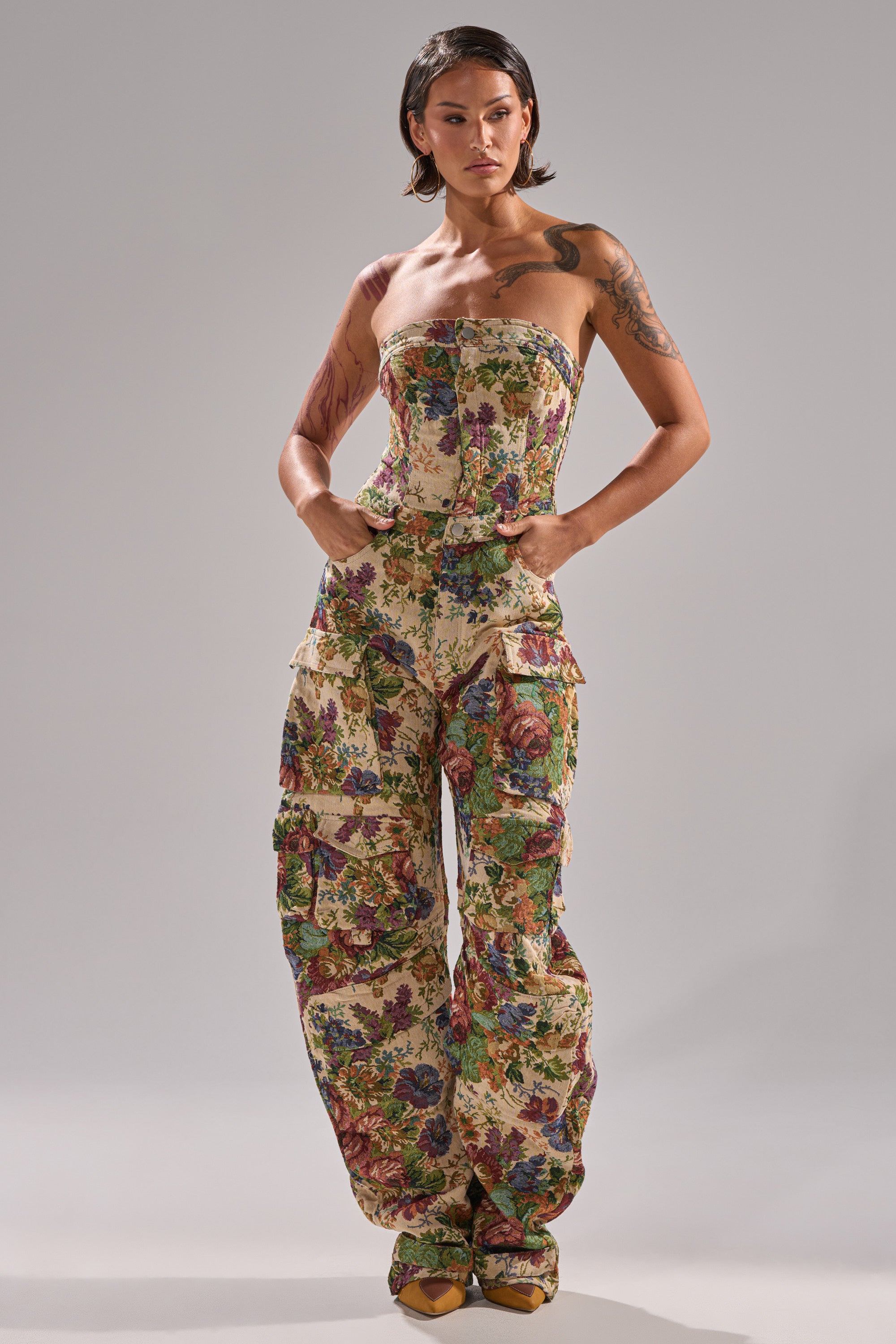 A woman with short dark hair and arm tattoos showcases effortless style in the IN HER ELEMENT FLORAL WOVEN JUMPSUIT, featuring a strapless cut, wide legs, and pockets, set against a plain gray background with yellow shoes.