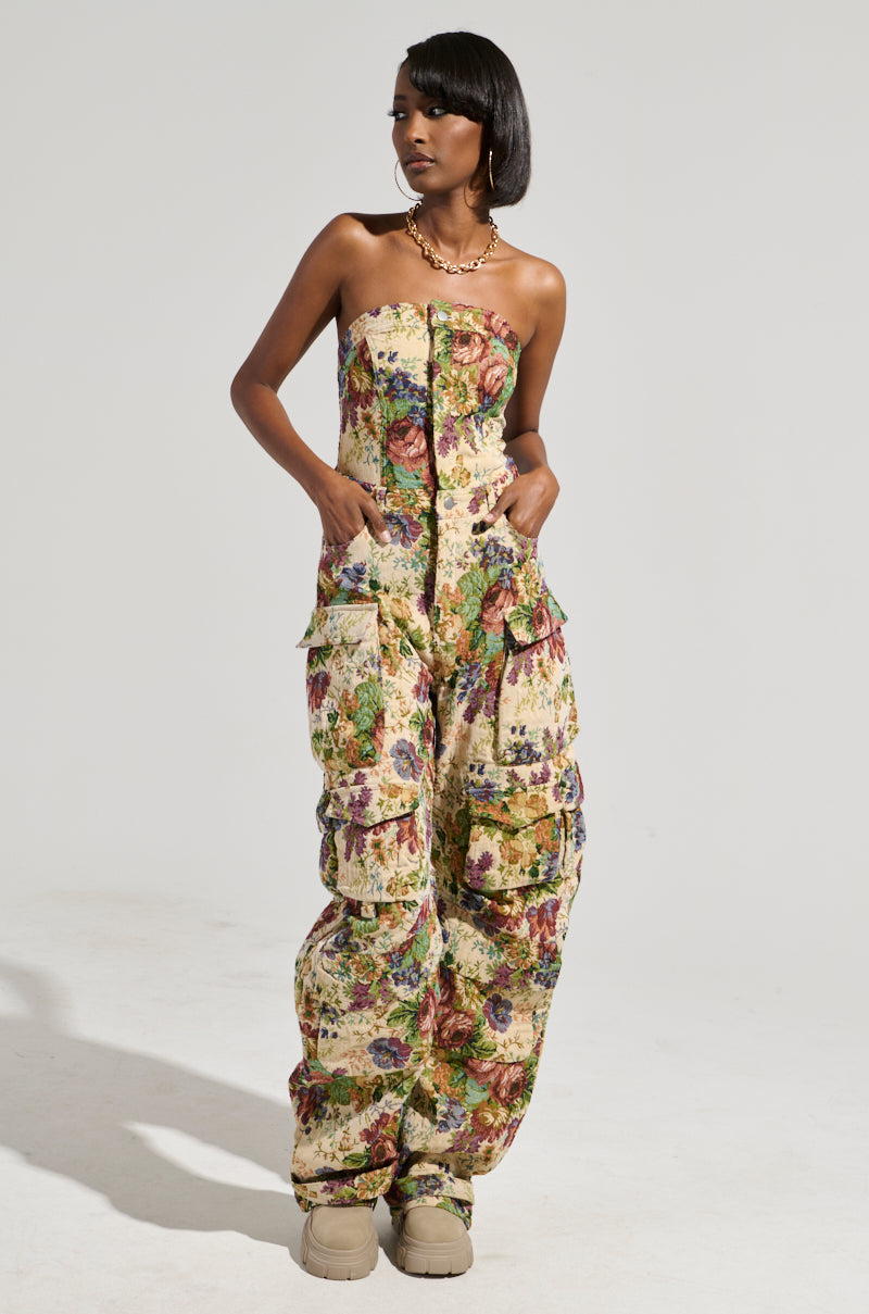 Wearing the IN HER ELEMENT FLORAL WOVEN JUMPSUIT featuring a strapless design, wide-leg silhouette, and multiple pockets, a person pairs this look with beige boots. Sporting short, dark hair and a necklace, they stand against a plain backdrop, gazing to the side with effortless style.