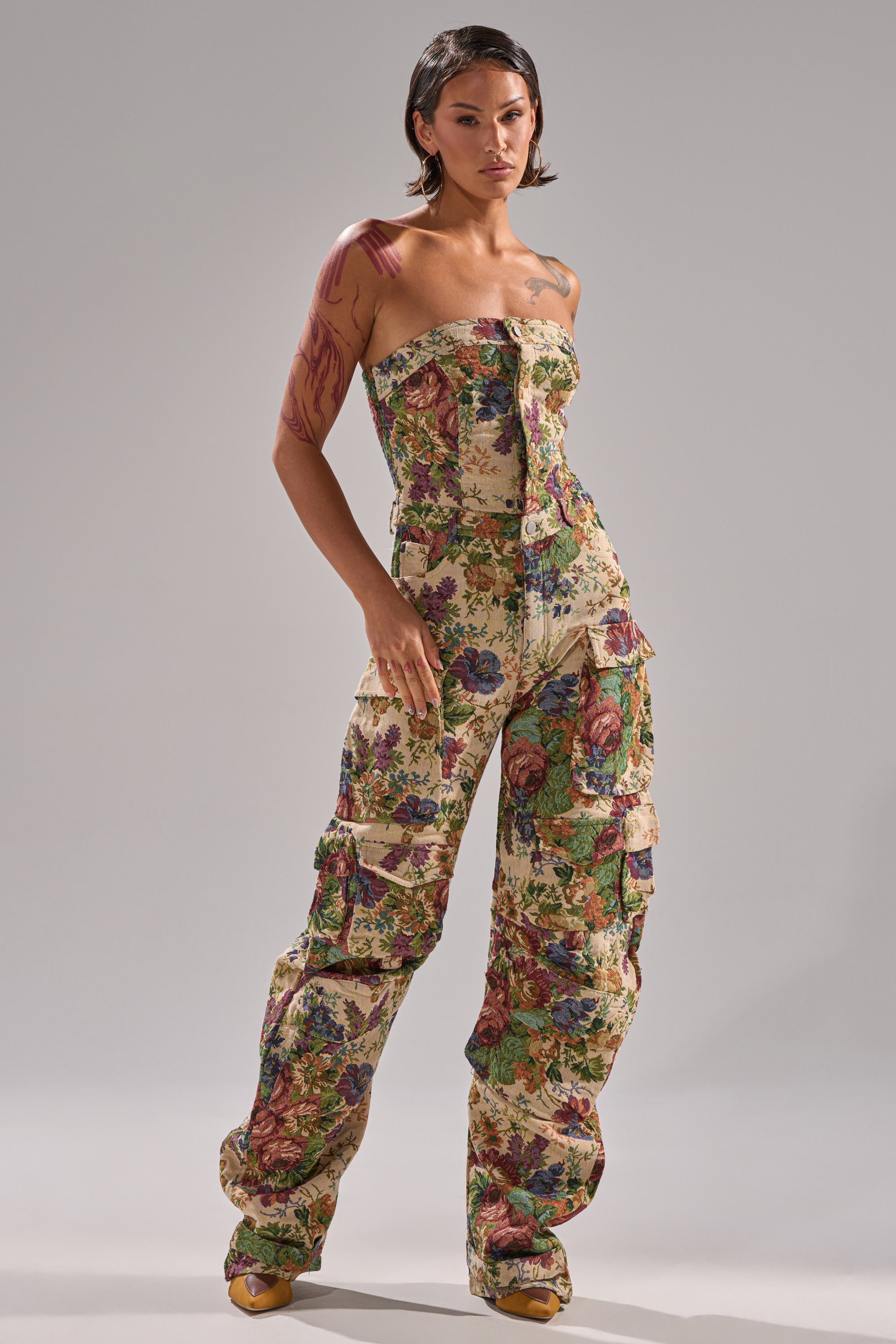 A person with short dark hair and arm tattoos models the IN HER ELEMENT FLORAL WOVEN JUMPSUIT, featuring a vibrant floral print, wide-leg cut, and multiple pockets, standing confidently against a plain gray background.