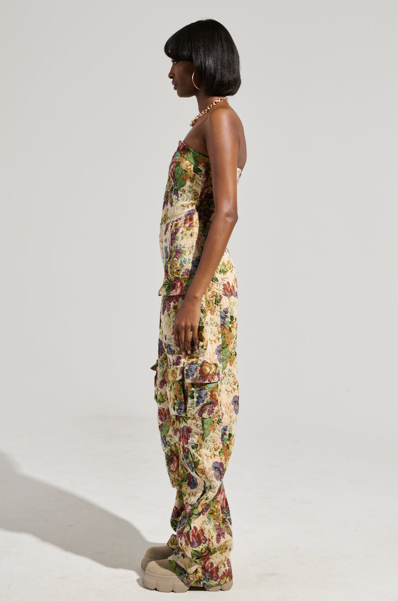 A person with short hair stands in profile, wearing the IN HER ELEMENT FLORAL WOVEN JUMPSUIT, which features a colorful and vintage floral pattern. The wide-leg silhouette contrasts elegantly against the plain, light background. Beige shoes complete the vibrant yet refined look.