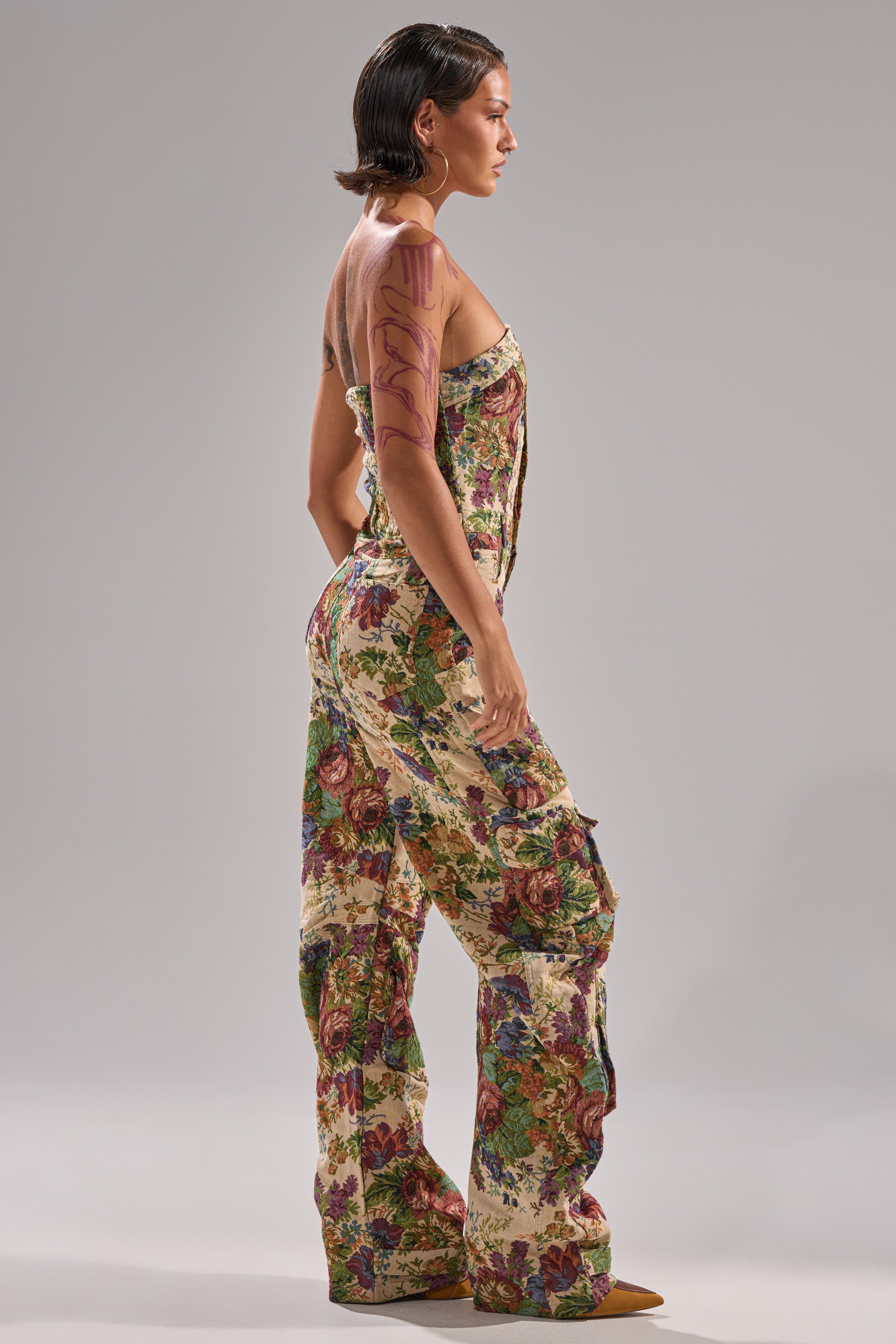 A person stands sideways in the IN HER ELEMENT FLORAL WOVEN JUMPSUIT, showcasing its sleeveless wide-leg design and bold floral print. Their short, slicked-back hair and abstract tattoos stand out against a plain gray background.
