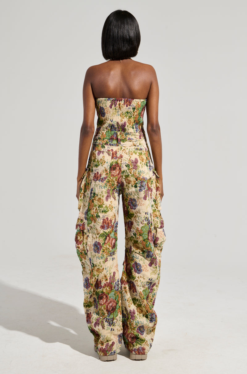 A person is standing with their back to the camera, donning the IN HER ELEMENT FLORAL WOVEN JUMPSUIT. This strapless piece boasts a wide leg silhouette and showcases vibrant colors along with an array of flowers, evoking a vintage floral aesthetic. The simple, neutral background highlights the standout nature of this clothing item.