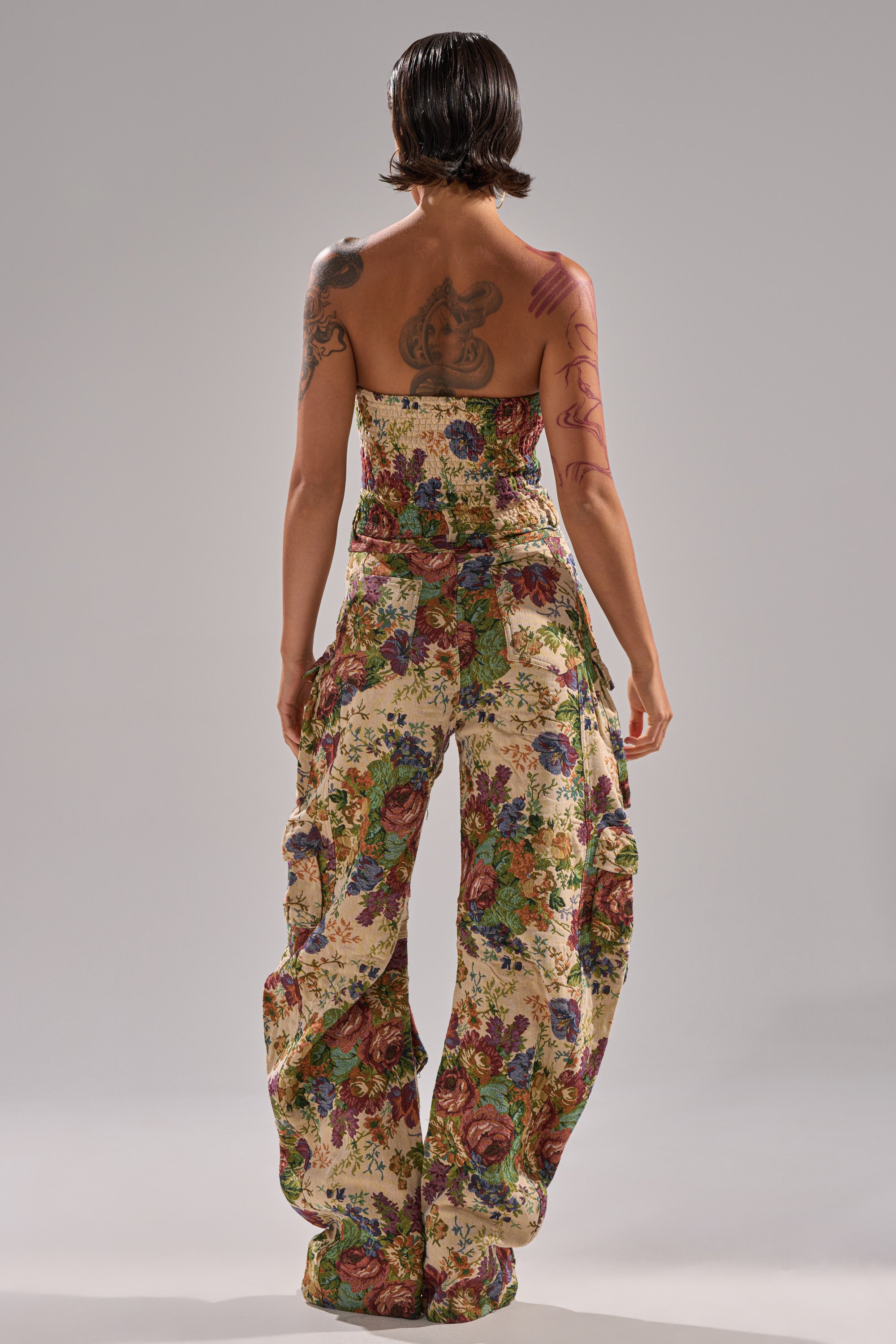 A tattooed person with short, wet-look hair stands with their back to the camera, wearing the IN HER ELEMENT FLORAL WOVEN JUMPSUIT, a strapless, wide-leg floral outfit, against a plain gray backdrop.