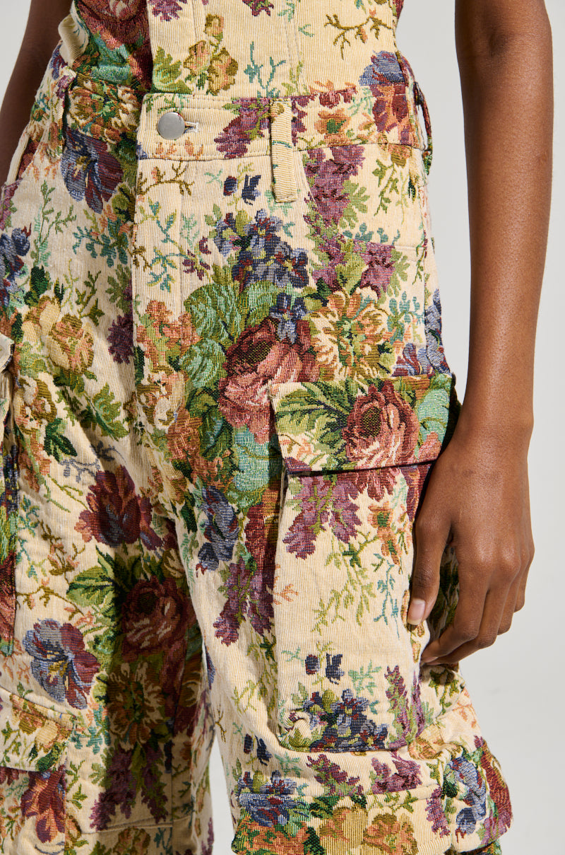 Close-up of a person wearing the IN HER ELEMENT FLORAL WOVEN JUMPSUIT, featuring a wide-leg silhouette and large pockets. The fabric displays colorful flowers in shades of red, purple, green, and blue on a light background. The person stands with their hand resting near a pocket.