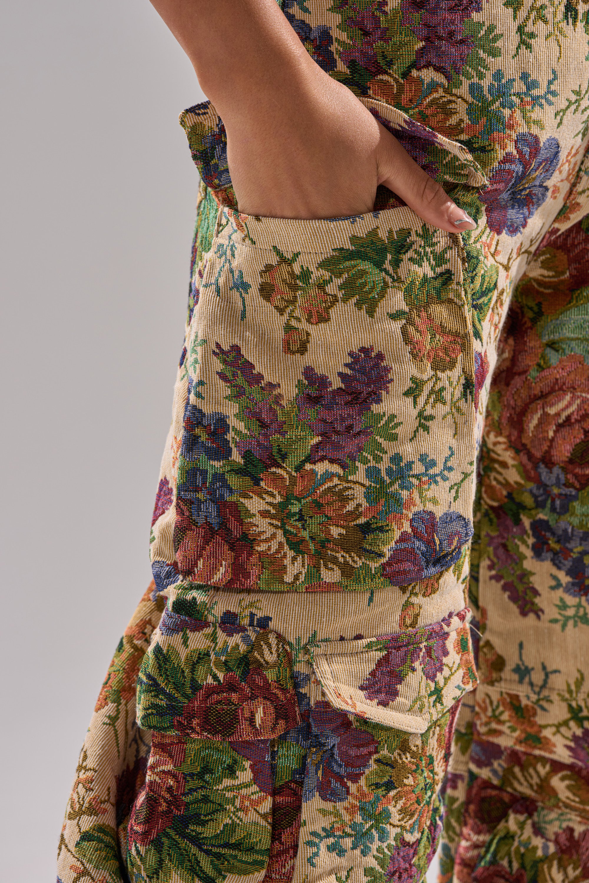 Close-up of a hand in the pocket of the IN HER ELEMENT FLORAL WOVEN JUMPSUIT, featuring detailed green, purple, red, and blue flowers on a beige background.