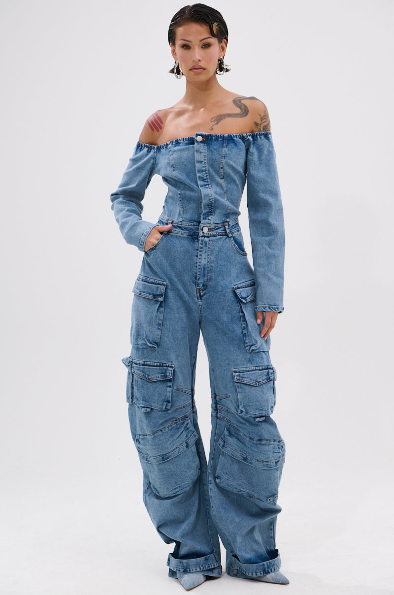 A person with short dark hair models the ARI LONG SLEEVE CARGO DENIM JUMPSUIT, featuring an off-the-shoulder design, large cargo pockets, and wide legs, against a plain white background.