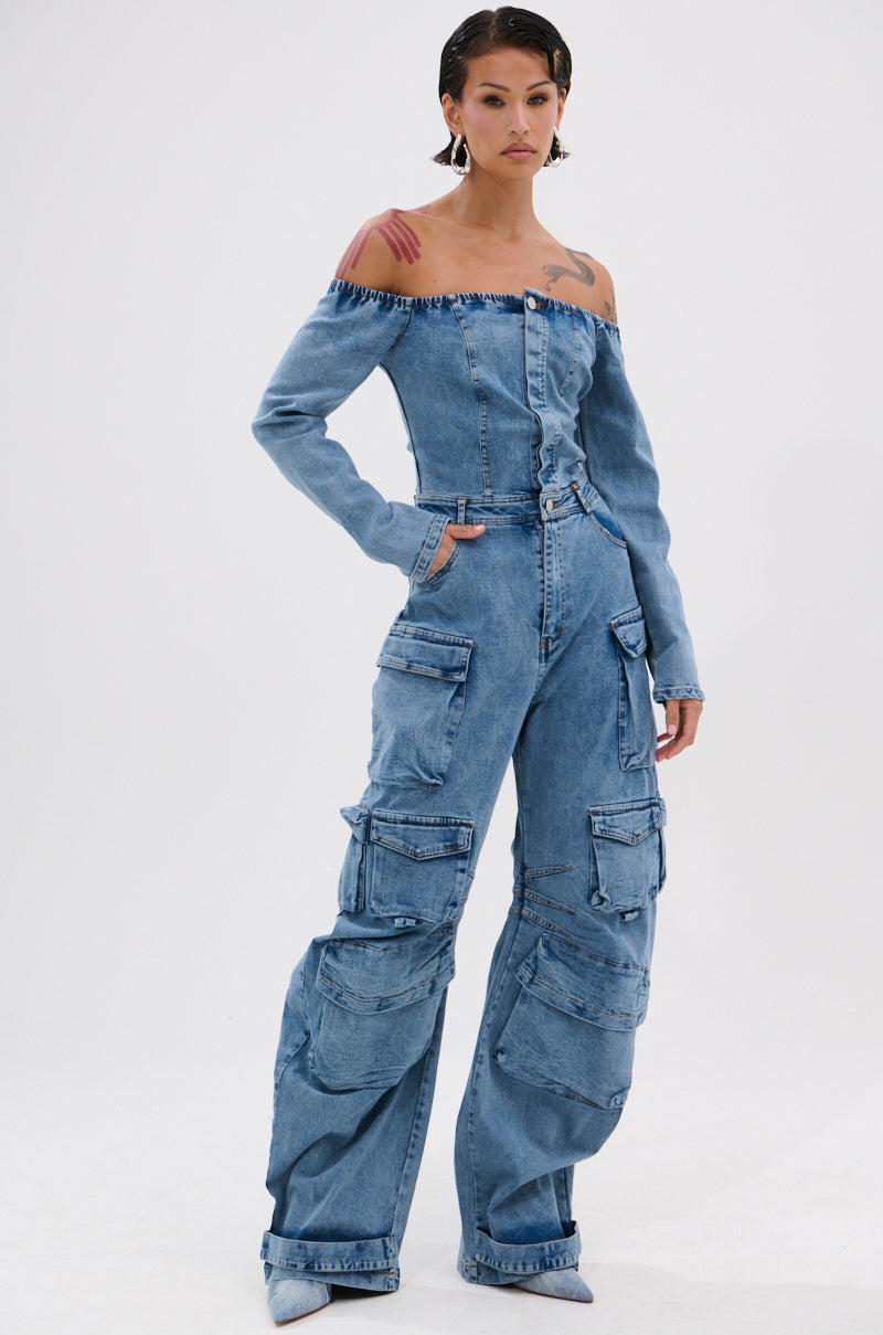Someone models the ARI LONG SLEEVE CARGO DENIM JUMPSUIT, featuring multiple cargo pockets and an off-the-shoulder design, while standing confidently against a plain white background.