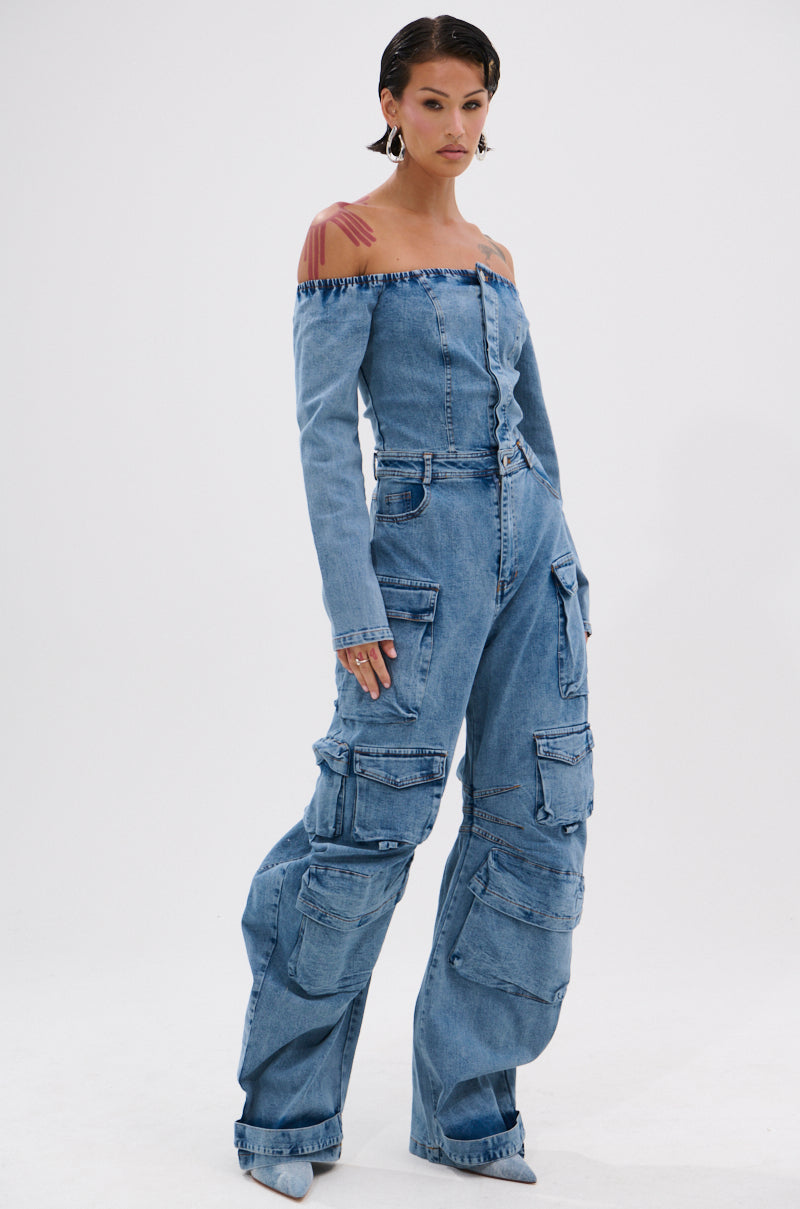 Wearing the ARI LONG SLEEVE CARGO DENIM JUMPSUIT with off-the-shoulder design and multiple cargo pockets, a person with slicked-back hair and pointed-toe shoes poses confidently against a plain white background.