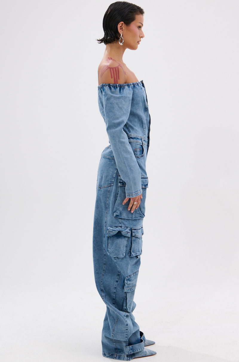 A woman models the ARI LONG SLEEVE CARGO DENIM JUMPSUIT, styled off-the-shoulder with cargo pockets. She stands in profile, wearing hoop earrings, pointed shoes, and has short hair with a trendy wet-look finish against a white background.