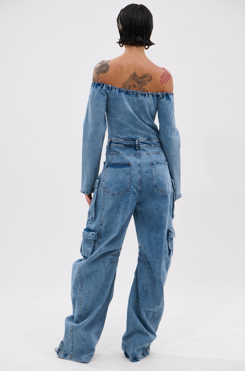 A person with short dark hair and tattoos wears the ARI LONG SLEEVE CARGO DENIM JUMPSUIT, standing with their back to the camera against a plain white background.