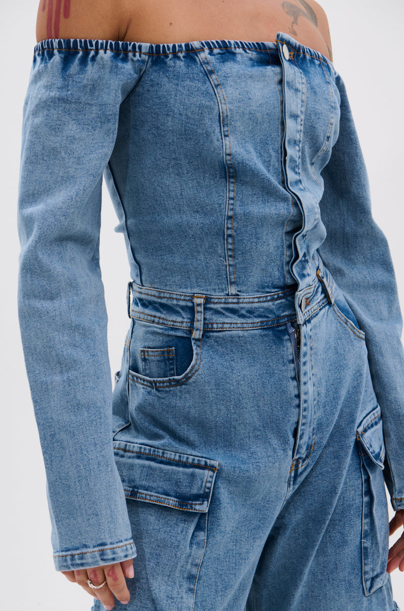 Wearing the ARI LONG SLEEVE CARGO DENIM JUMPSUIT, a person shows off its light blue denim, chic cargo pockets on the top and pants, long sleeves, off-the-shoulder design, and fitted waist.