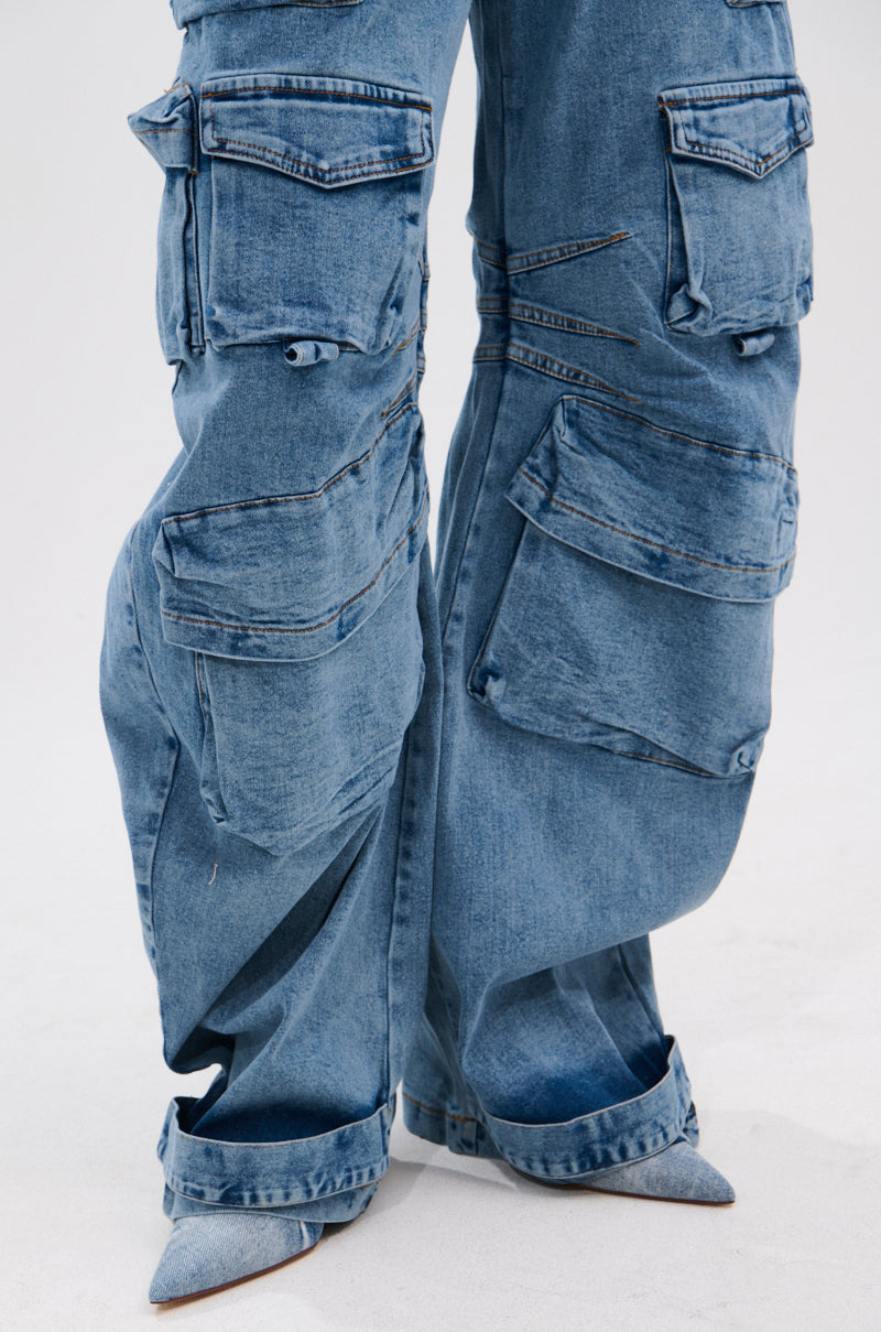 Close-up of the ARI LONG SLEEVE CARGO DENIM JUMPSUIT in blue, featuring multiple cargo pockets, seam details, and a loose wide-leg fit. Styled with pointed-toe shoes against a plain light background.