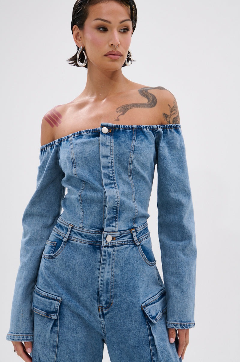 A woman with short dark hair wears the ARI LONG SLEEVE CARGO DENIM JUMPSUIT, featuring an off-the-shoulder design and cargo pockets. She accessorizes with large hoop earrings and neutral makeup, posing against a plain white background.