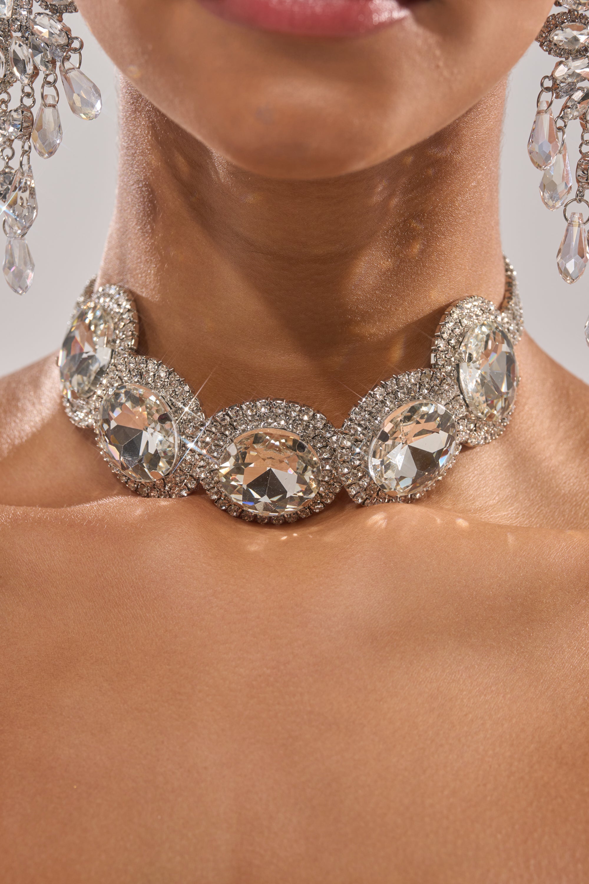 A close-up of a woman's neck wearing the AT THE BALL CHOKER, an evening accessory with large oval crystals framed by rhinestones, paired with matching crystal earrings visible on the sides. 1
