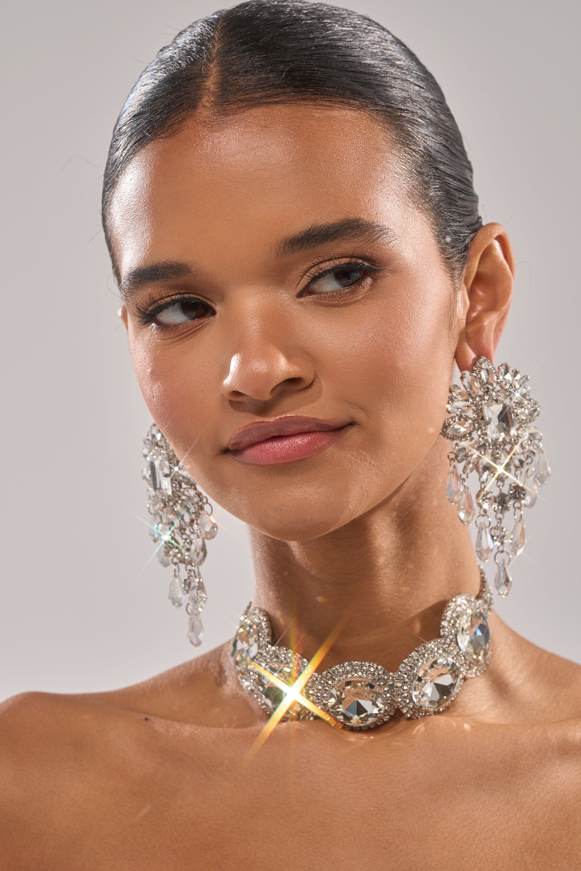 A woman with smooth, dark hair pulled back wears the AT THE BALL CHOKER—a sparkling rhinestone accessory—paired with ornate crystal earrings. Her natural makeup and calm, confident look highlight this elegant eveningwear piece. 2