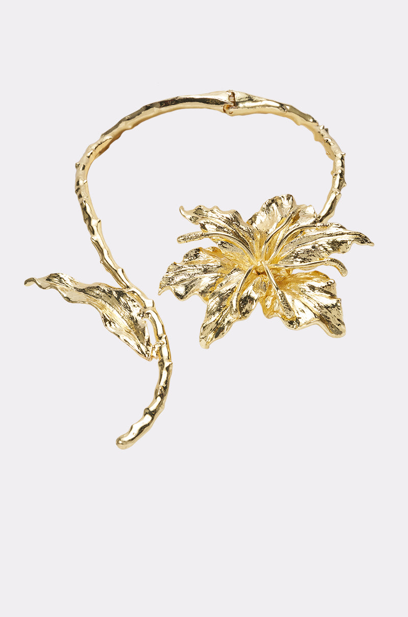 The EXTRA IS MY NAME NECKLACE is a gold floral choker adorned with a large, textured flower and leaf motif, featuring intricate detailing on both the petals and leaves—an elegant and sophisticated blooming flower necklace.