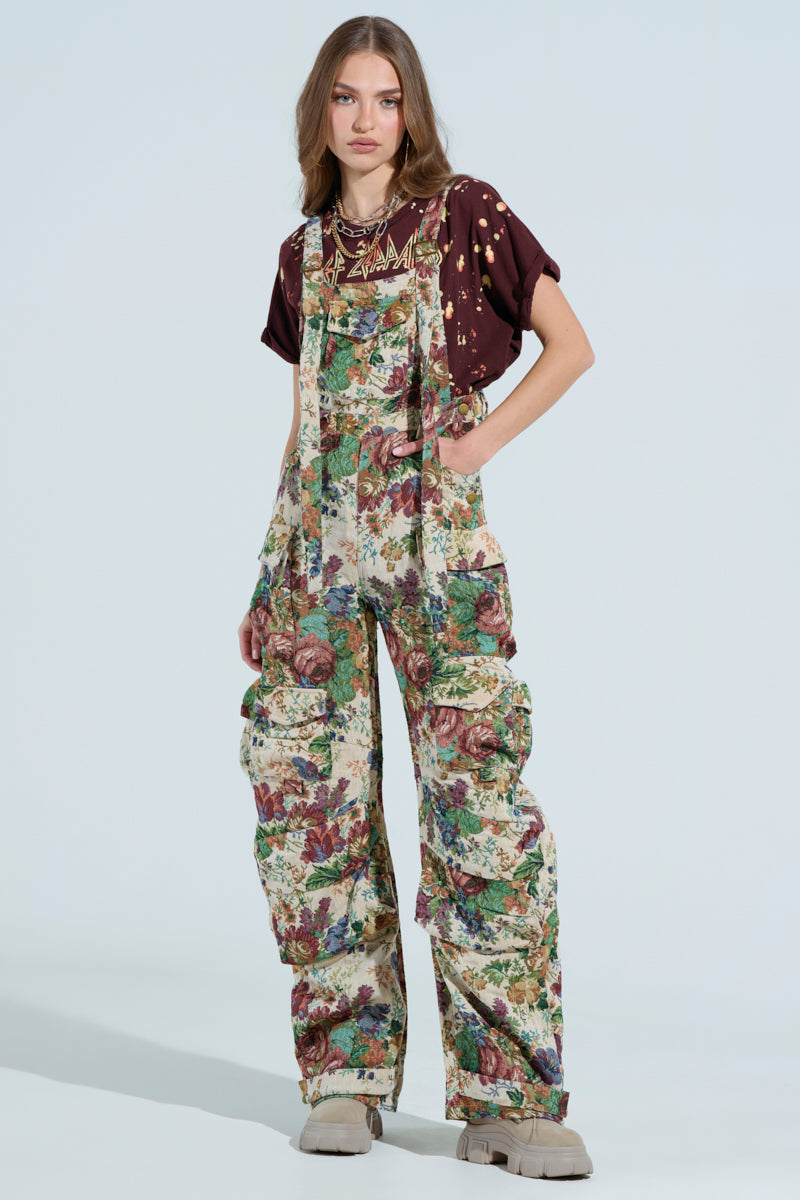 Wearing AKIRA Label's LIFE THESE DAYS OVERALLS featuring a wide-leg floral print, they pair it with a dark T-shirt adorned with gold accents. Their long hair flows freely, perfectly matched by light-colored chunky shoes against a neutral background.
