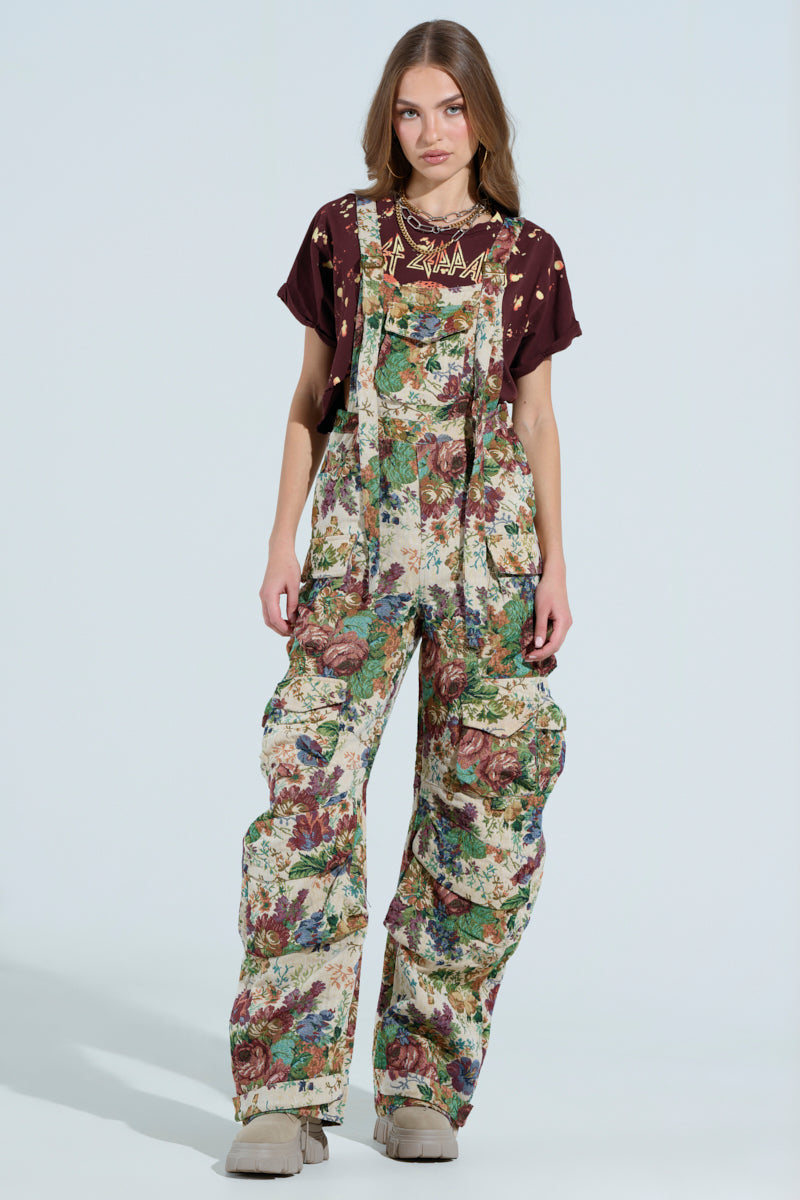 A person is wearing LIFE THESE DAYS floral-print overalls with a burgundy graphic T-shirt. The loose, wide-leg design is complemented by beige boots, creating a vibrant contrast against the plain white background.
