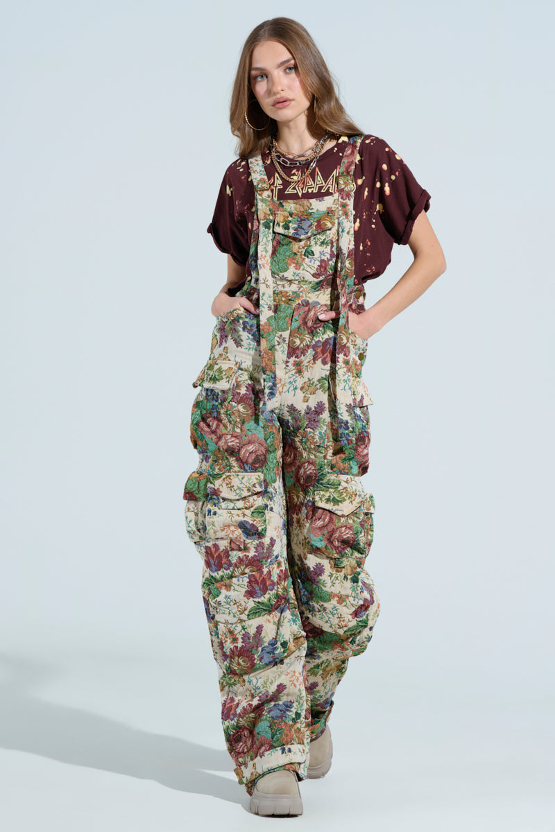 Someone is showcasing AKIRA Label's LIFE THESE DAYS OVERALLS with a wide-leg silhouette in green, red, and beige floral print. They are paired effortlessly with a brown printed T-shirt against a plain, light-colored background.