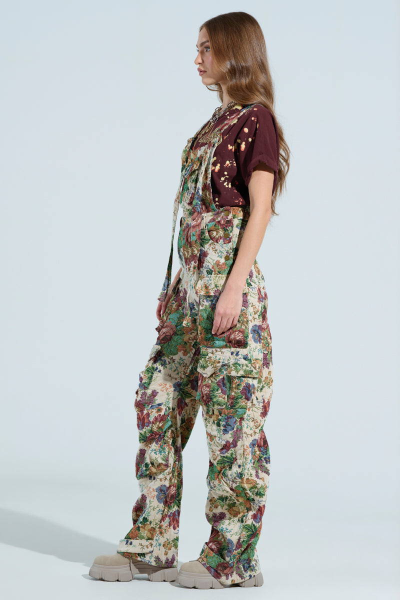A woman with long brown hair stands in profile against a light background, wearing a dark brown t-shirt and colorful LIFE THESE DAYS OVERALLS in green, purple, and cream hues with beige shoes. The chic overalls feature a wide leg silhouette.