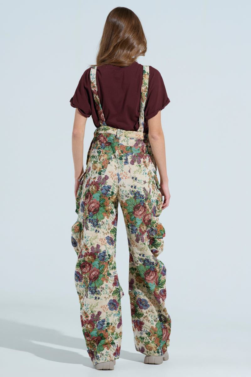 A person stands with their back to the camera in floral print overalls from the AKIRA Label, known as the LIFE THESE DAYS OVERALLS, with a brown shirt and long hair down. They're wearing chunky shoes against a plain light blue background.