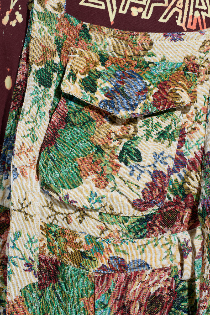 Close-up of the LIFE THESE DAYS OVERALLS showcasing a floral tapestry pattern in green, red, and purple. Features include a button-flap pocket with zigzag stitching and an upper part displaying partial logo letters.