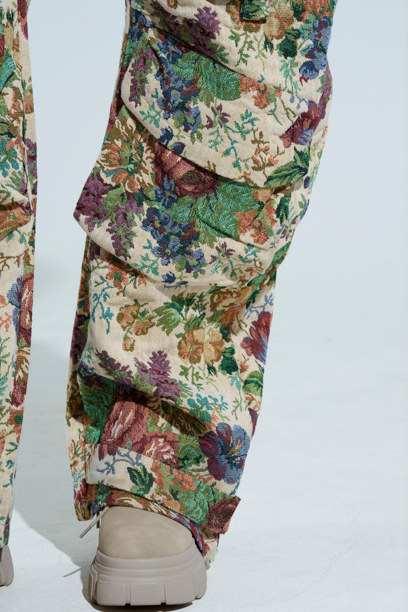 Close-up of a person wearing LIFE THESE DAYS OVERALLS by AKIRA Label, showcasing a multicolored floral tapestry pattern. The wide leg silhouette features vibrant green, purple, red, and blue flowers on a cream background, paired with light beige shoes.