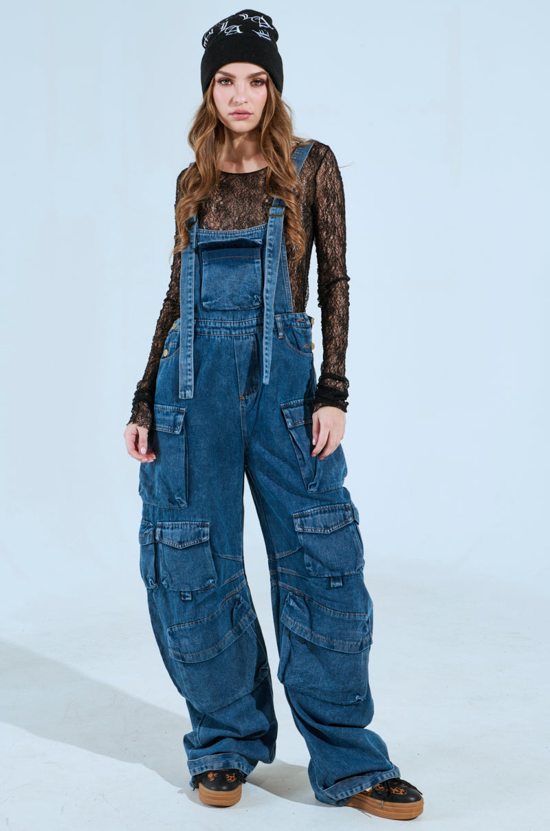 Wearing the ALWAYS ON YOUR MIND DENIM JUMPSUIT IN BLUE, featuring adjustable straps, a medium wash, and paired with a black lace top, chunky shoes, and a black beanie, a person with long wavy hair stands neutrally against a plain backdrop.