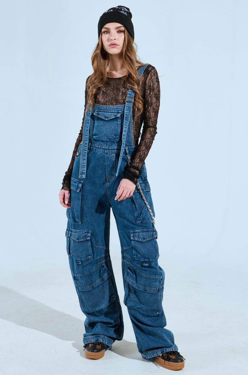 A person confidently stands with long hair, hands in pockets, wearing a black beanie, sheer black top, and the "Always On Your Mind Denim Jumpsuit in Blue" featuring wide-leg, medium-wash denim with multiple pockets, posed against a light background.