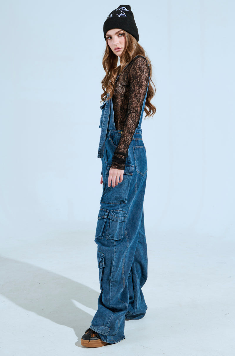 In a studio setting with a light background, a person confidently poses wearing the "Always On Your Mind Denim Jumpsuit in Blue" paired with a black lace top, brown shoes, and a black beanie.