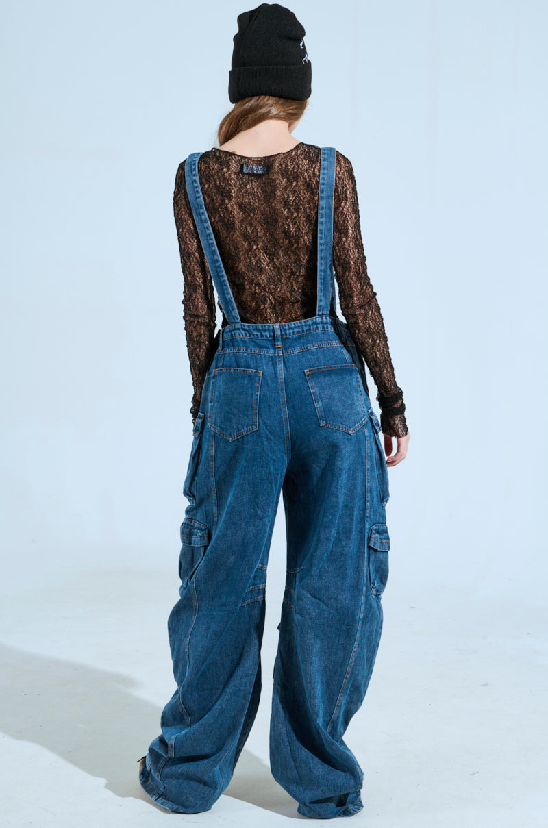 A person wearing a black lace top, a black beanie, and the ALWAYS ON YOUR MIND DENIM JUMPSUIT IN BLUE is standing with their back to the camera. The jumpsuit features wide legs, adjustable straps, and large side pockets against a plain light background.